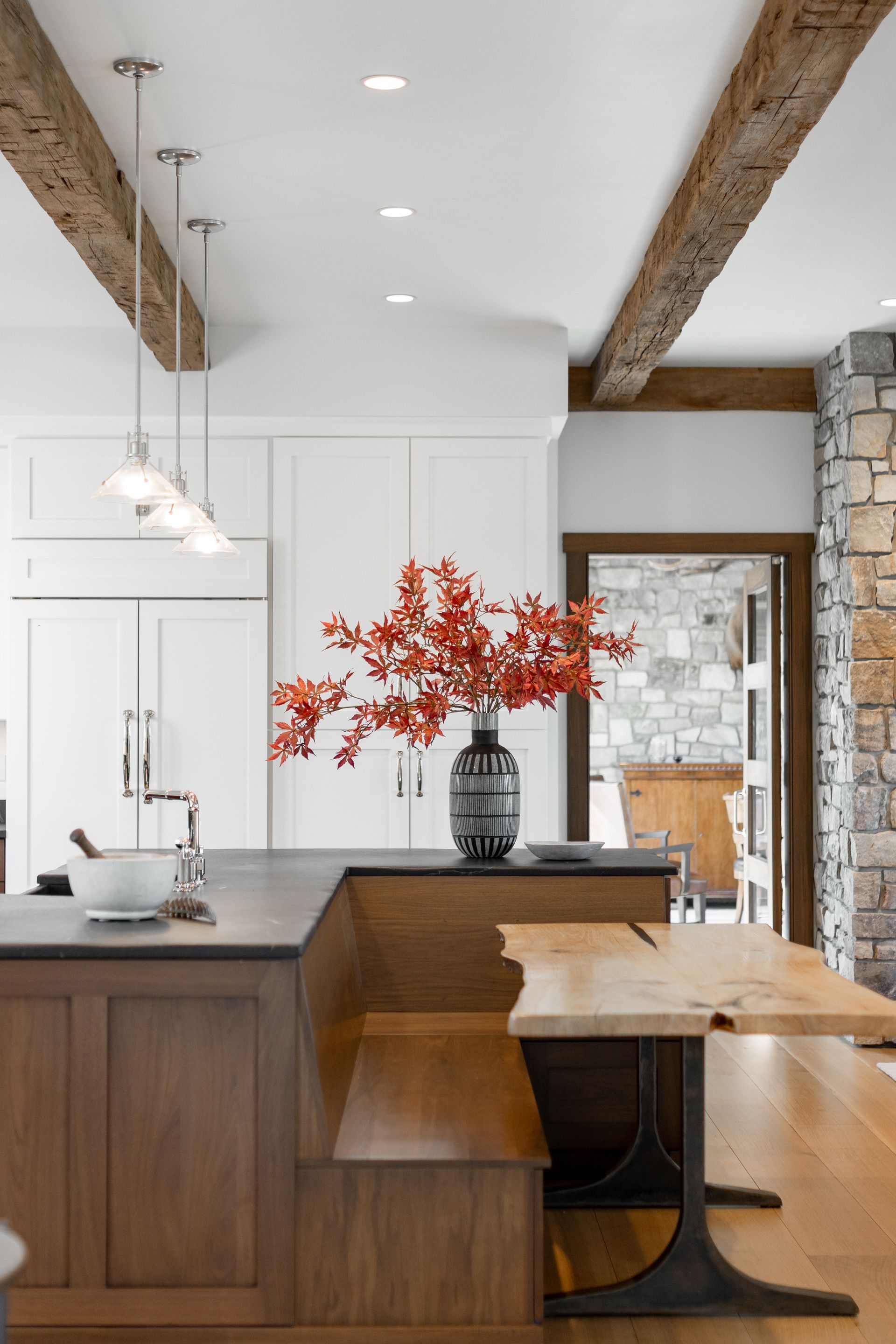 A wooden kitchen island with a built-in booth, live-edge dining table, and pendant lights under rustic ceiling beams.