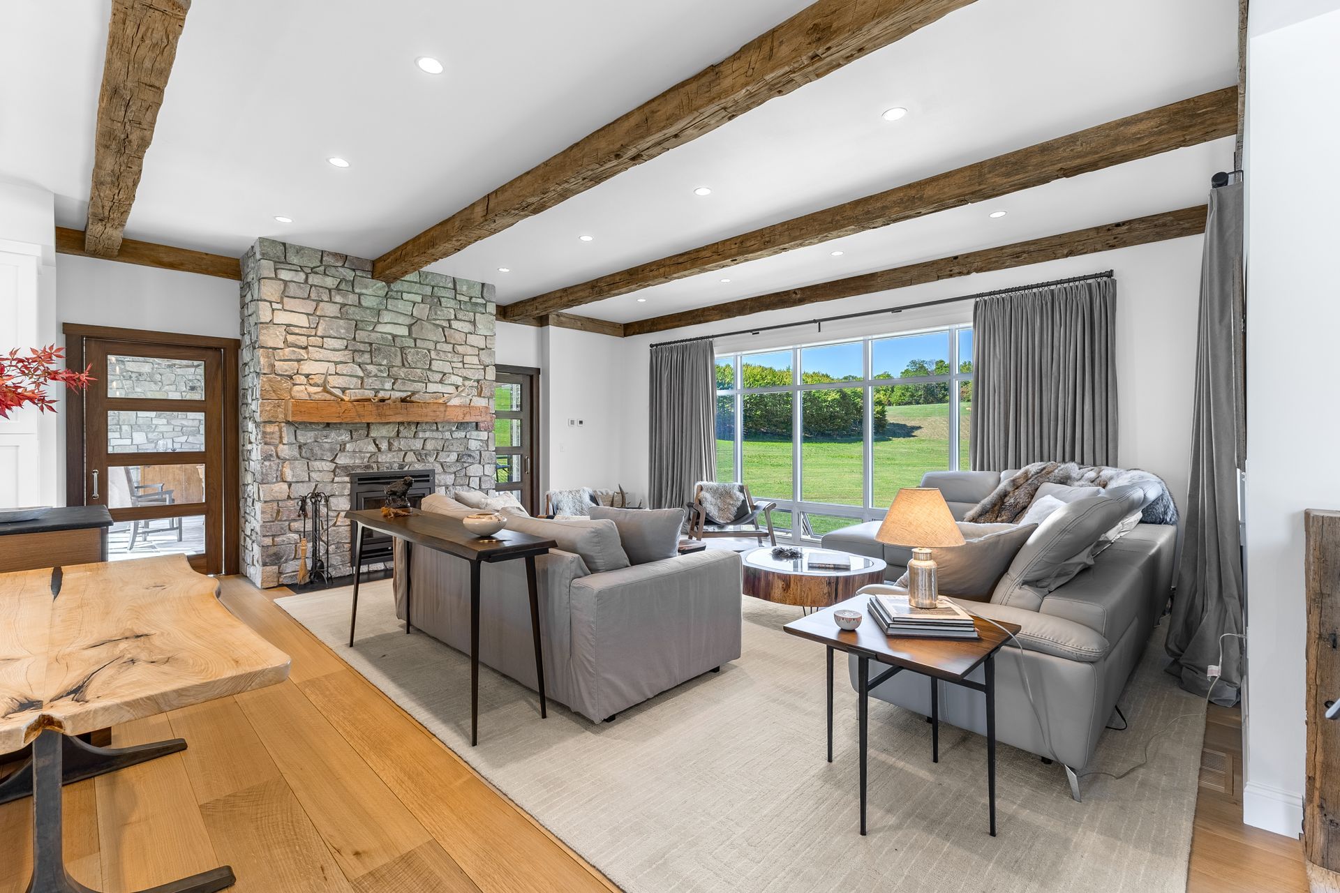 A cozy living room with wooden ceiling beams, a stone fireplace, grey sofas, and large windows looking out to a green field.