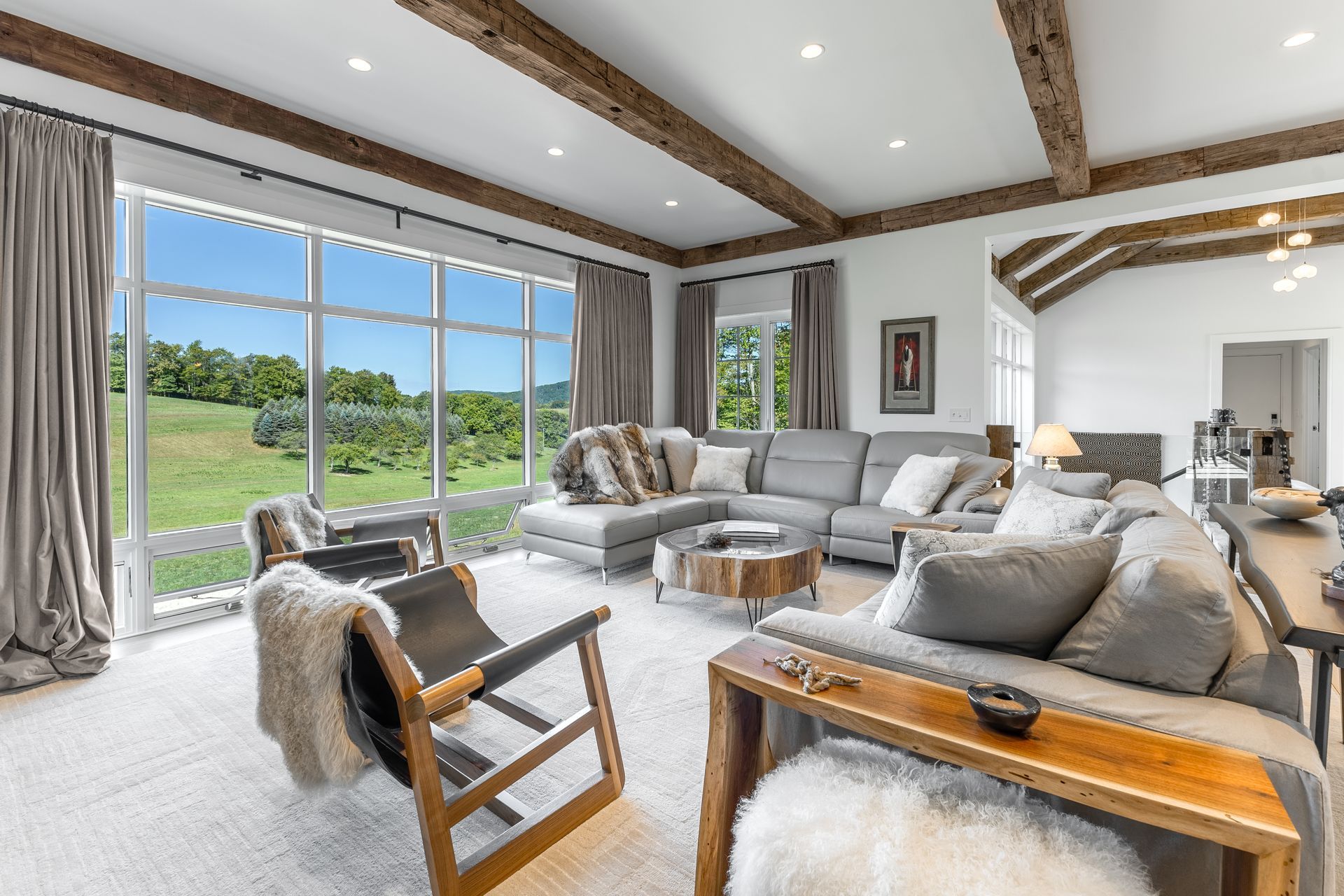 A bright living room features a sectional sofa, two wooden armchairs, ceiling beams, and a large window with rural views.