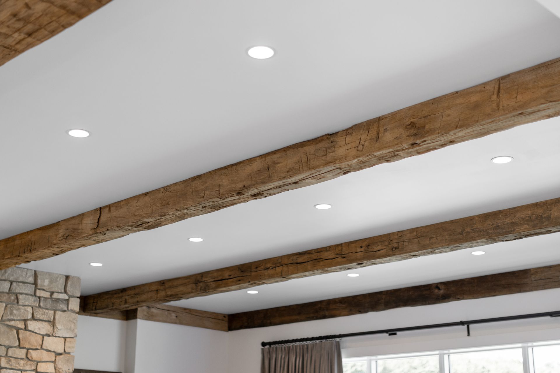 White ceiling with recessed lighting and decorative rustic wooden beams above a stone wall and window.