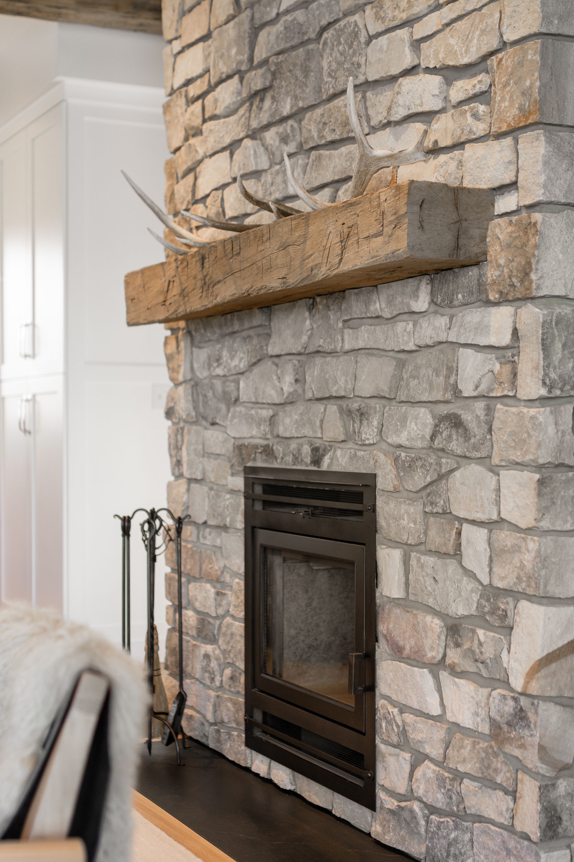 A stone fireplace with a thick, rustic wooden mantel decorated with antlers, featuring a black metal firebox and tools.