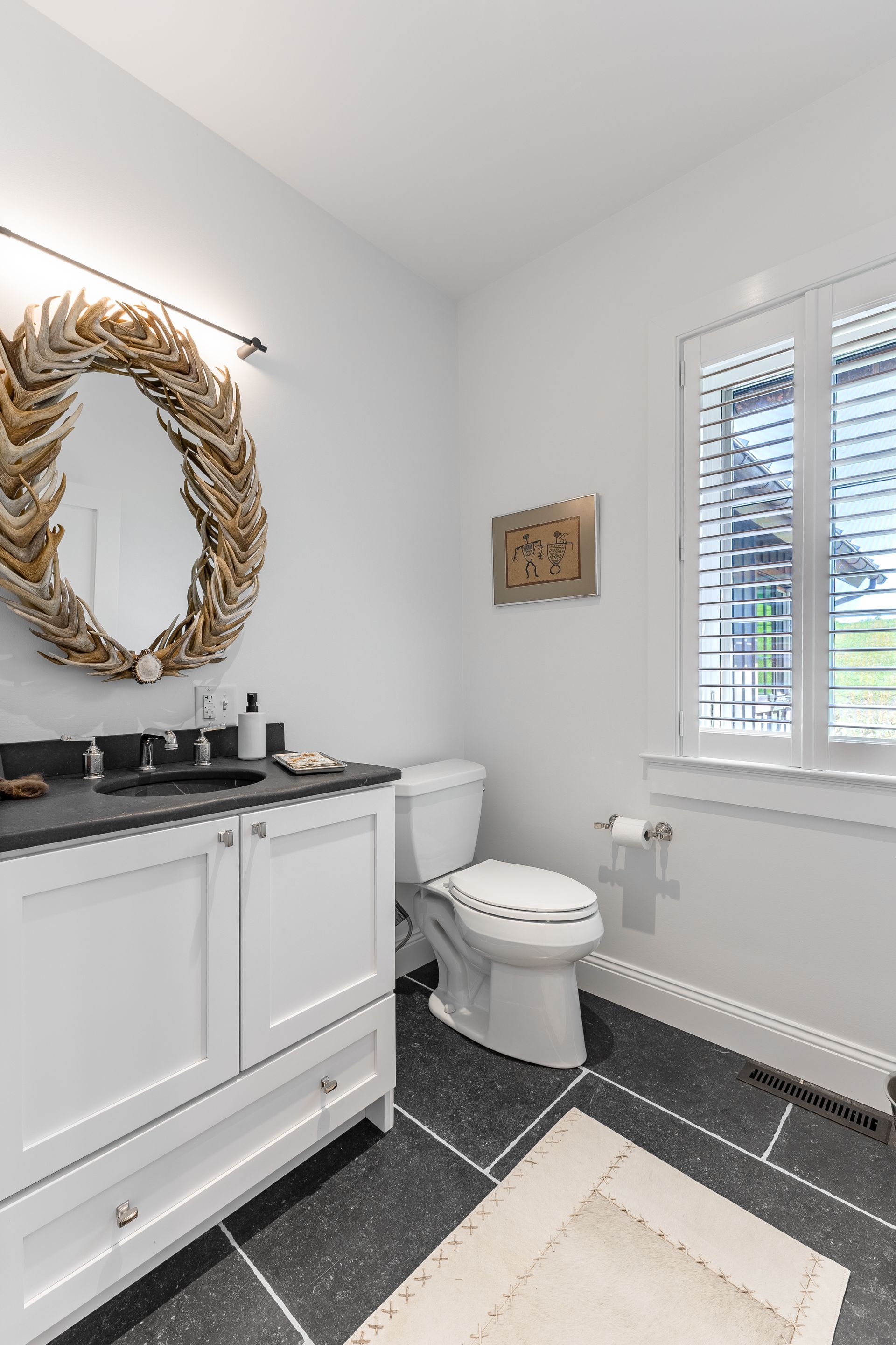 A bathroom with a white vanity, a dark countertop, an ornate antler mirror, a toilet, and a window with white shutters.