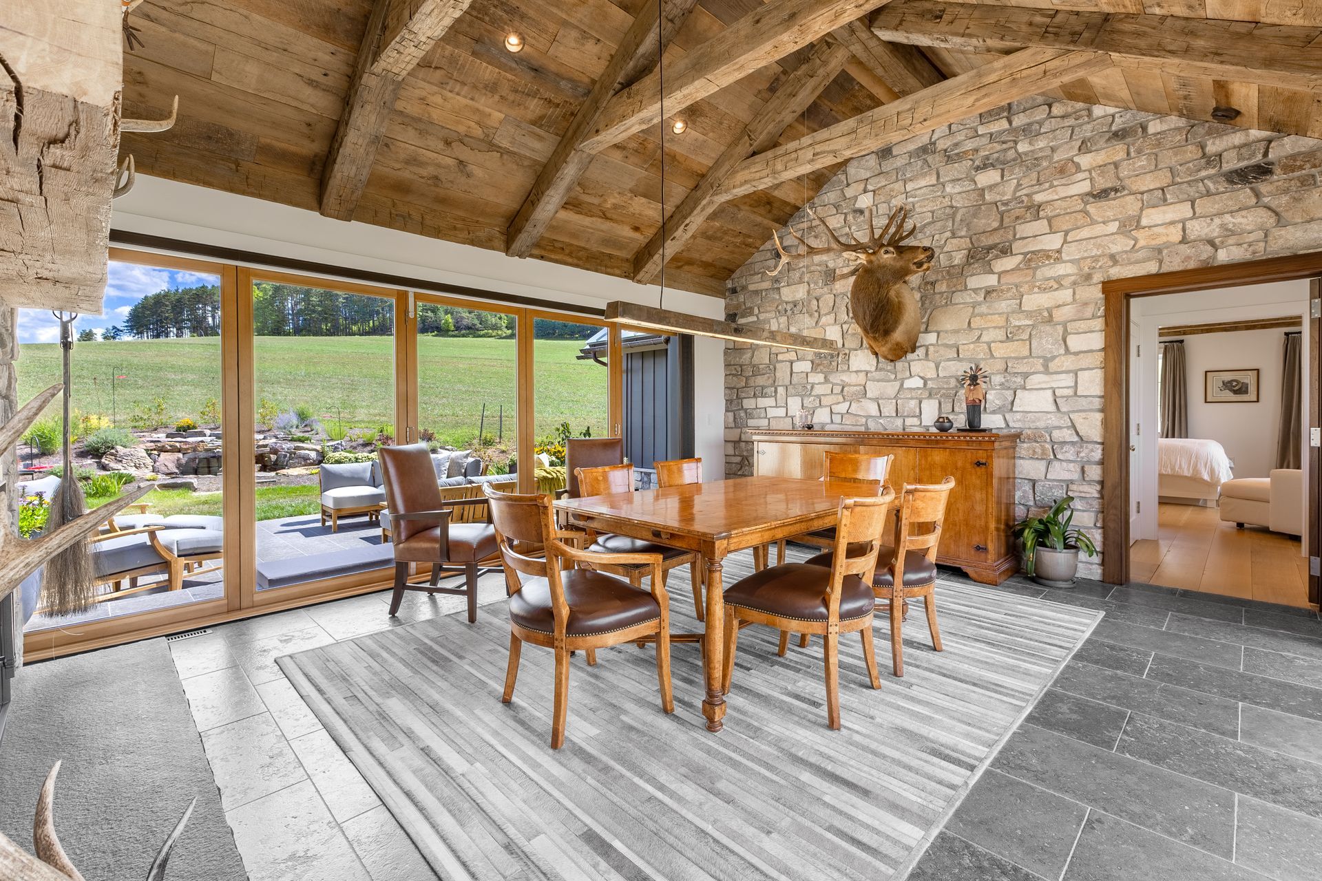 A rustic dining room with a wooden table, chairs, stone walls, exposed beams, and large windows overlooking a green field.