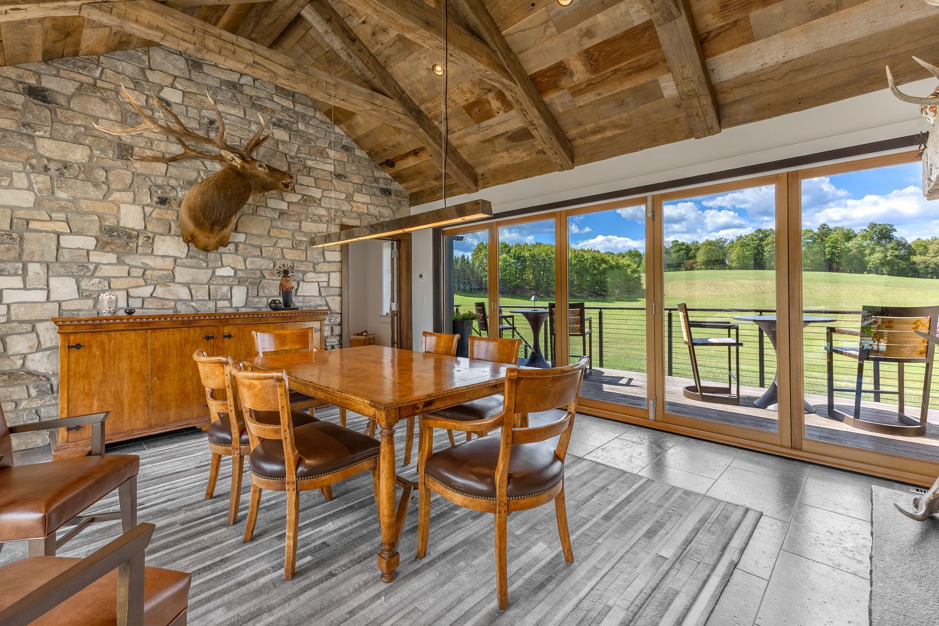 A rustic dining area with a stone wall, wooden table, leather chairs, and large glass doors overlooking a green field.