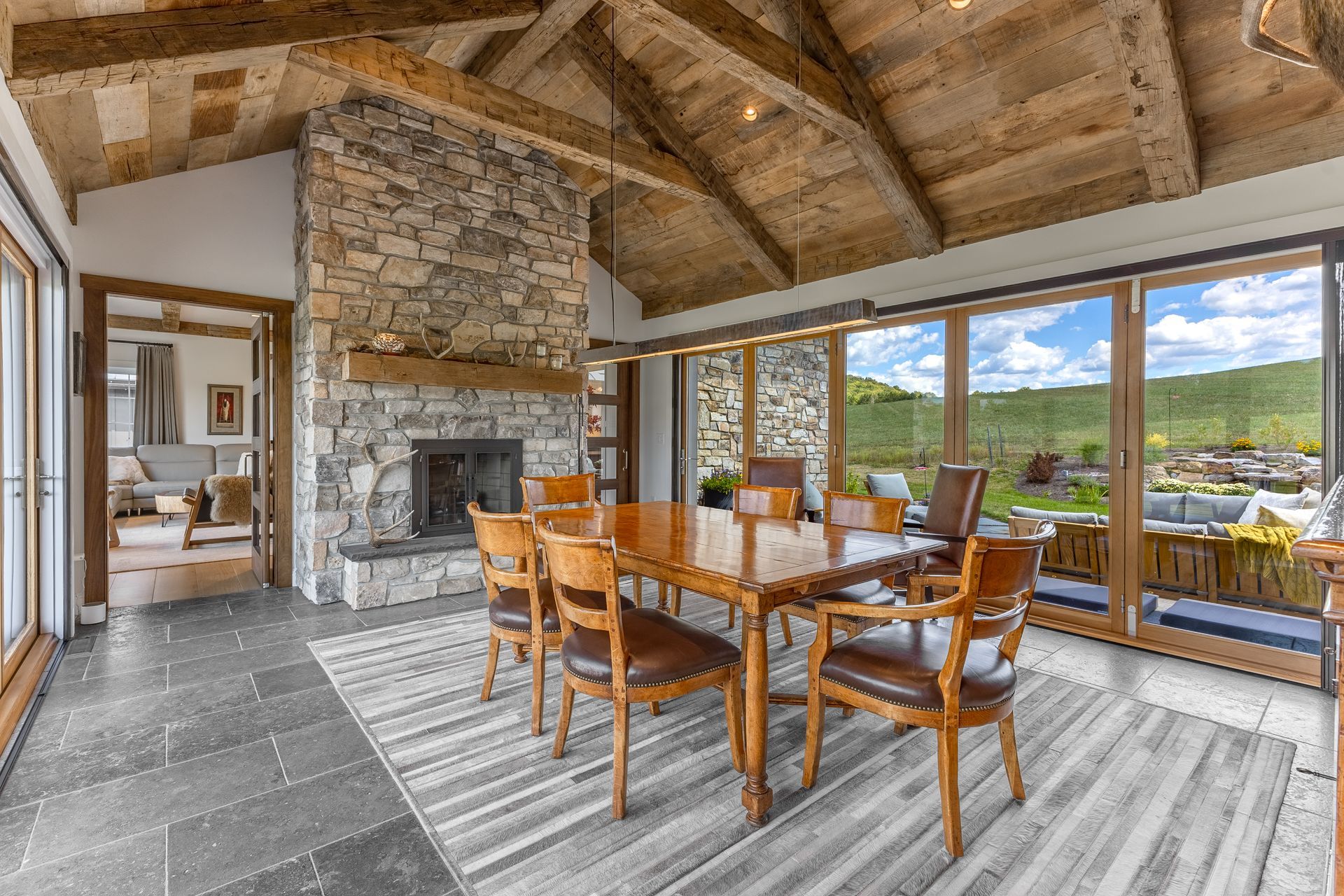 A rustic dining room with a stone fireplace, exposed wooden ceiling beams, large windows, and a table with six chairs.