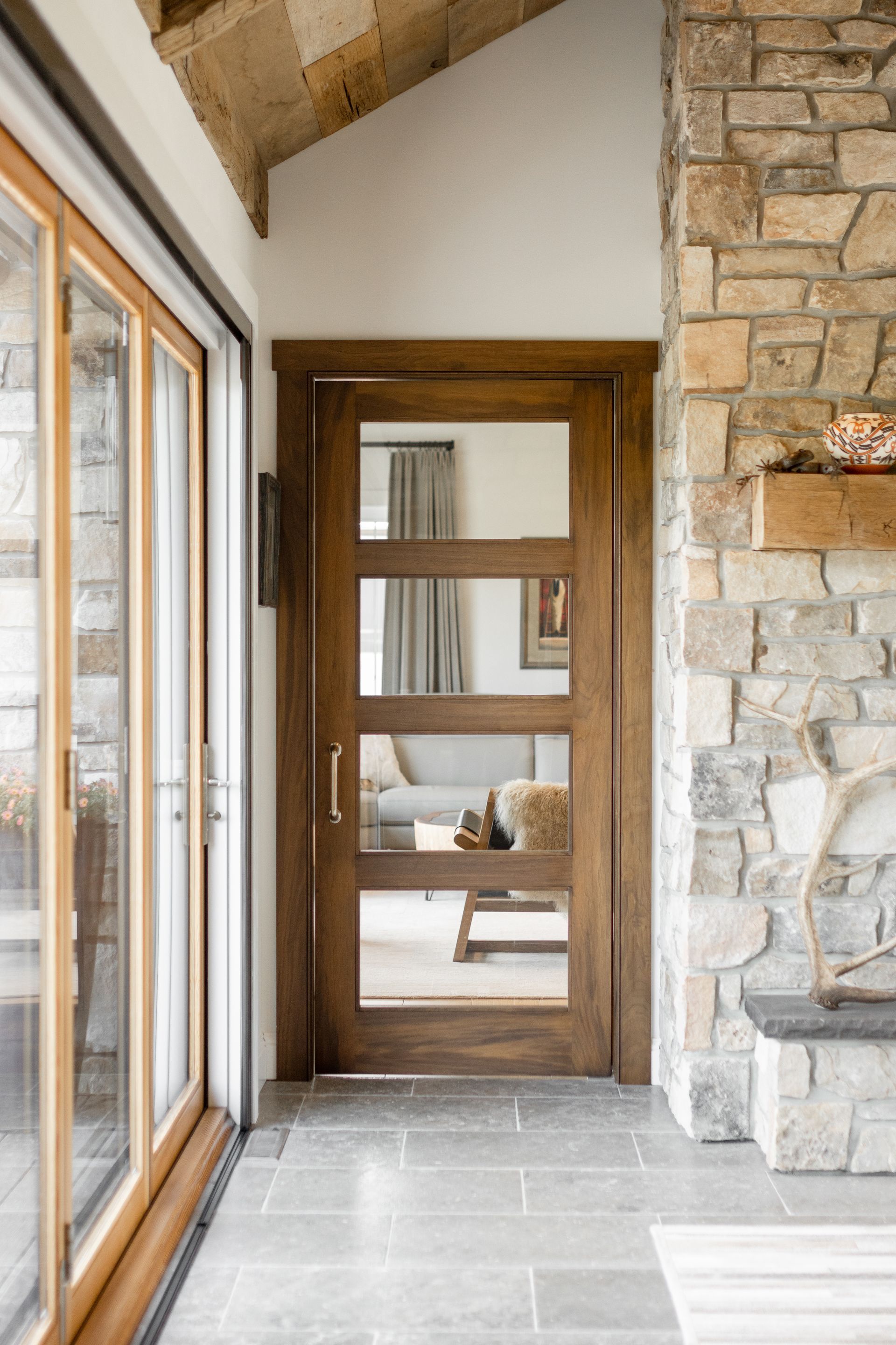 Interior view of a rustic hall with a stone wall, slate floor, and a wooden, four-pane glass door leading to a room.