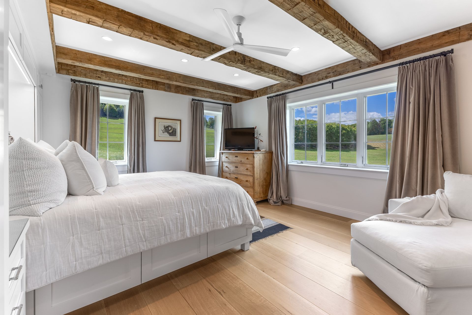 Bright bedroom with a king bed, wooden ceiling beams, large windows overlooking a green landscape, and white furniture.