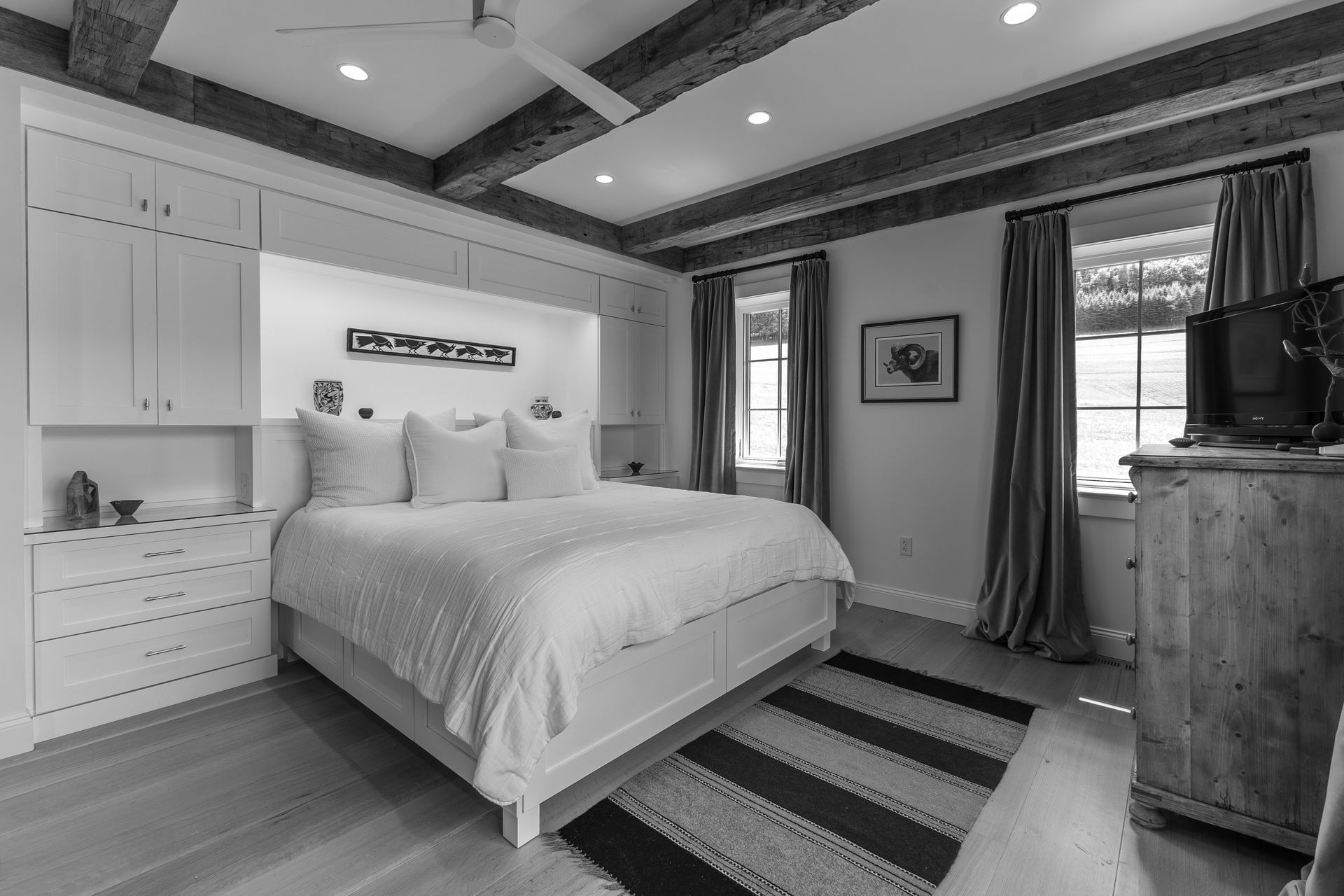 A bedroom with exposed ceiling beams, a white bed, built-in cabinets, a patterned rug, and a TV on a wooden dresser.