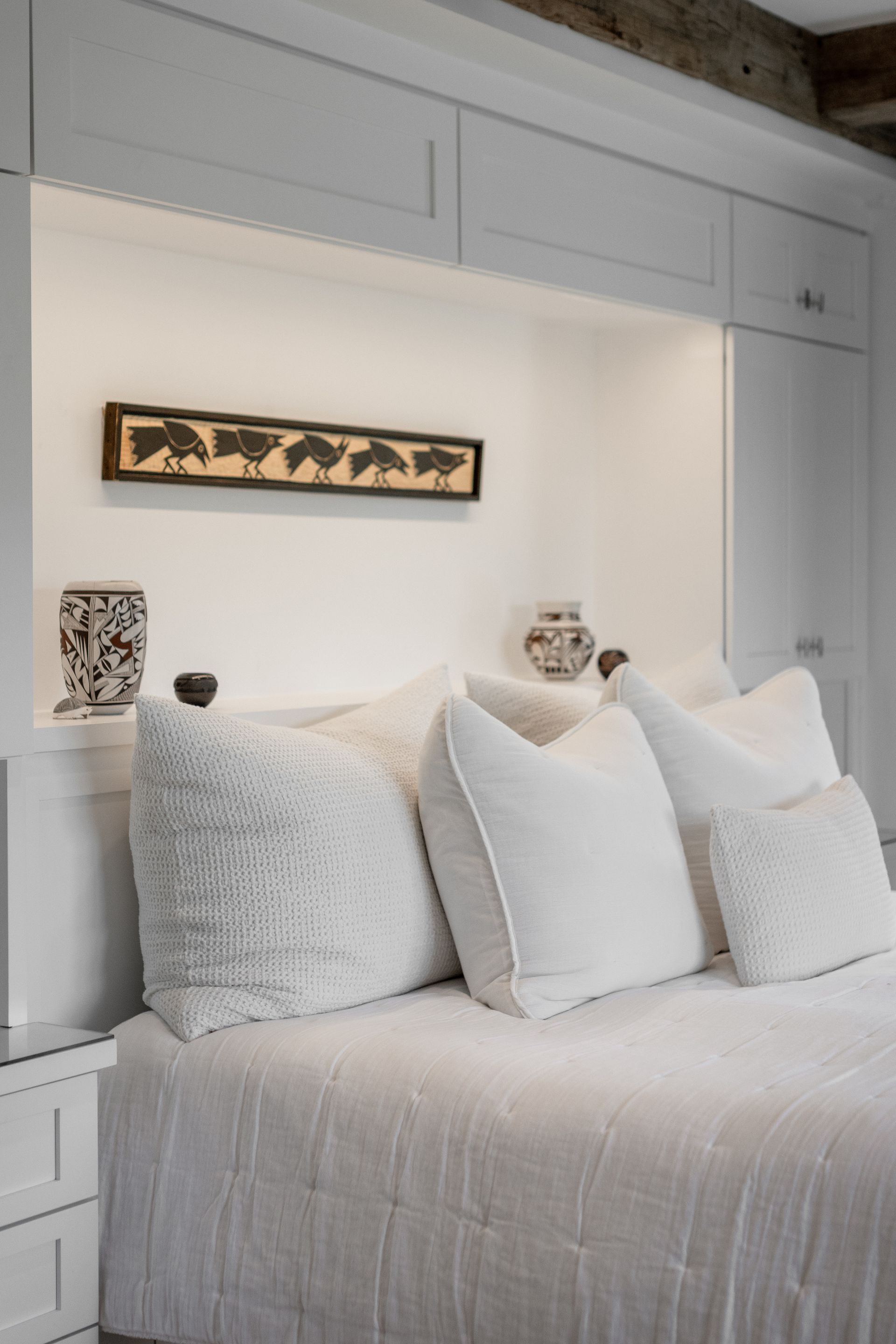 A white bedroom with a bed featuring textured pillows, under a recessed shelf displaying a long piece of tribal art.