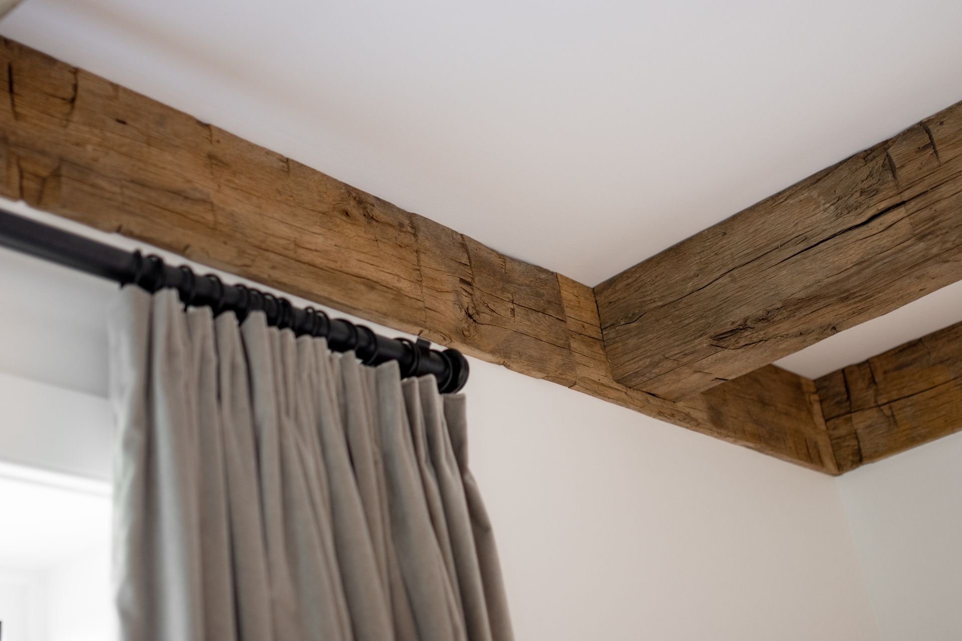 A close-up of heavy, rustic wooden ceiling beams meeting a white ceiling above a window with gray pleated curtains.