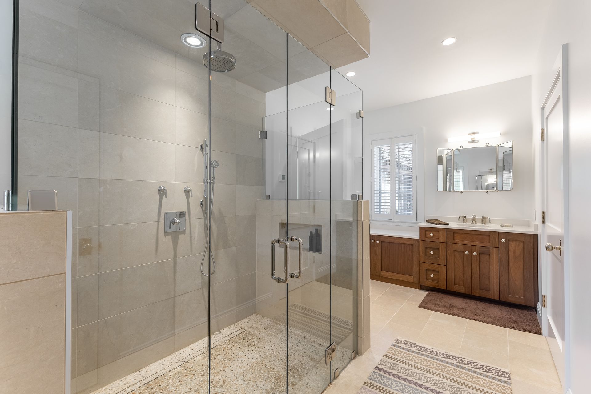 A modern bathroom featuring a large glass walk-in shower with pebble-stone flooring next to a wooden vanity and mirror.
