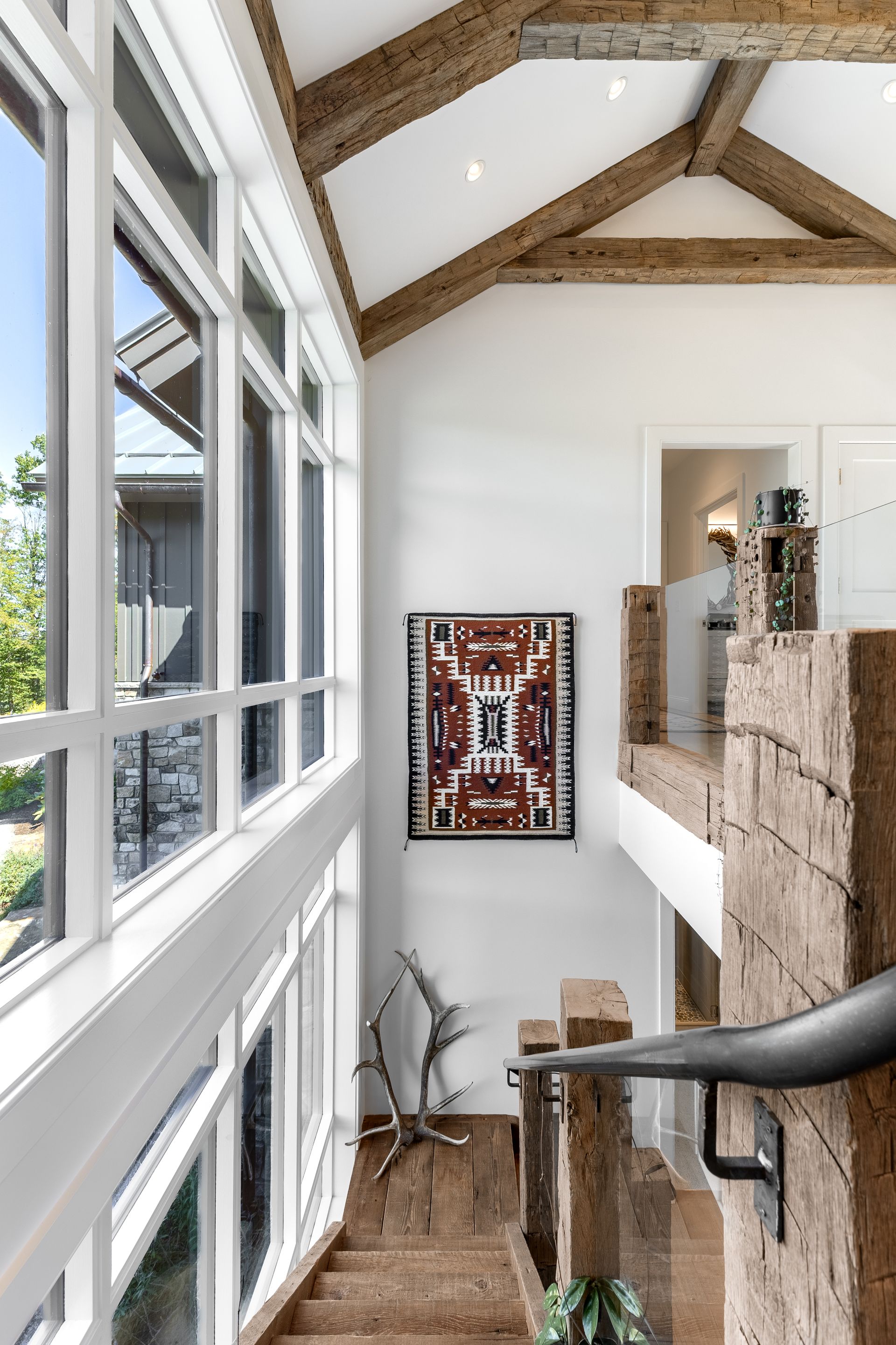 A wooden staircase leads up to a landing with a geometric rug on the wall, under vaulted ceilings with exposed beams.
