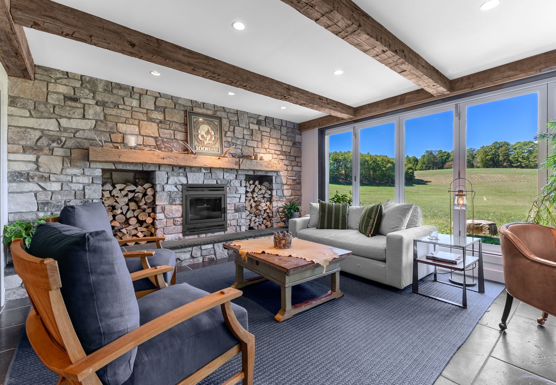 Modern living room with stone fireplace, gray sofa, blue chairs, and large windows overlooking fields
