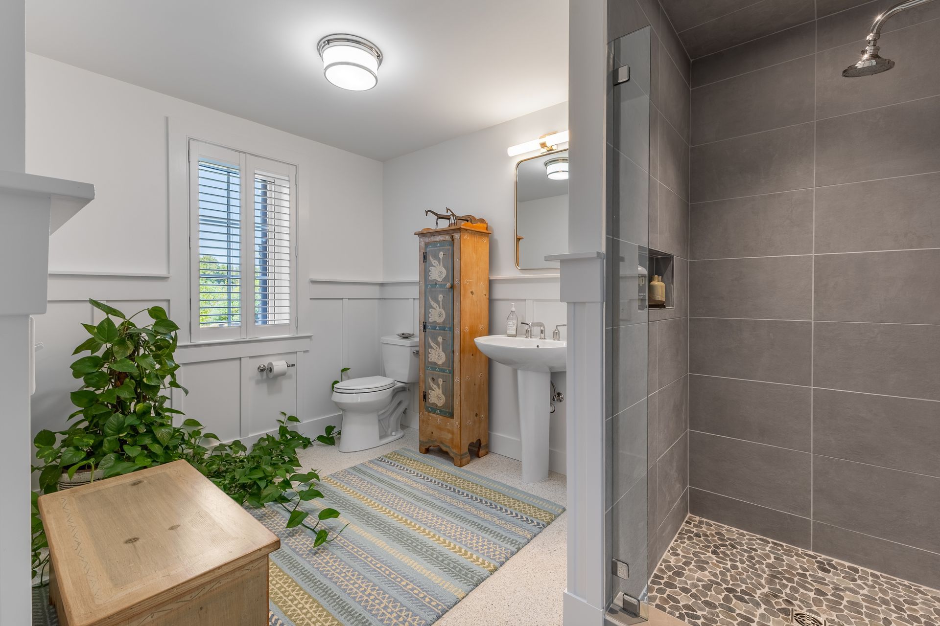 Bright bathroom with white fixtures, a plant, patterned rug, and a glass shower enclosure