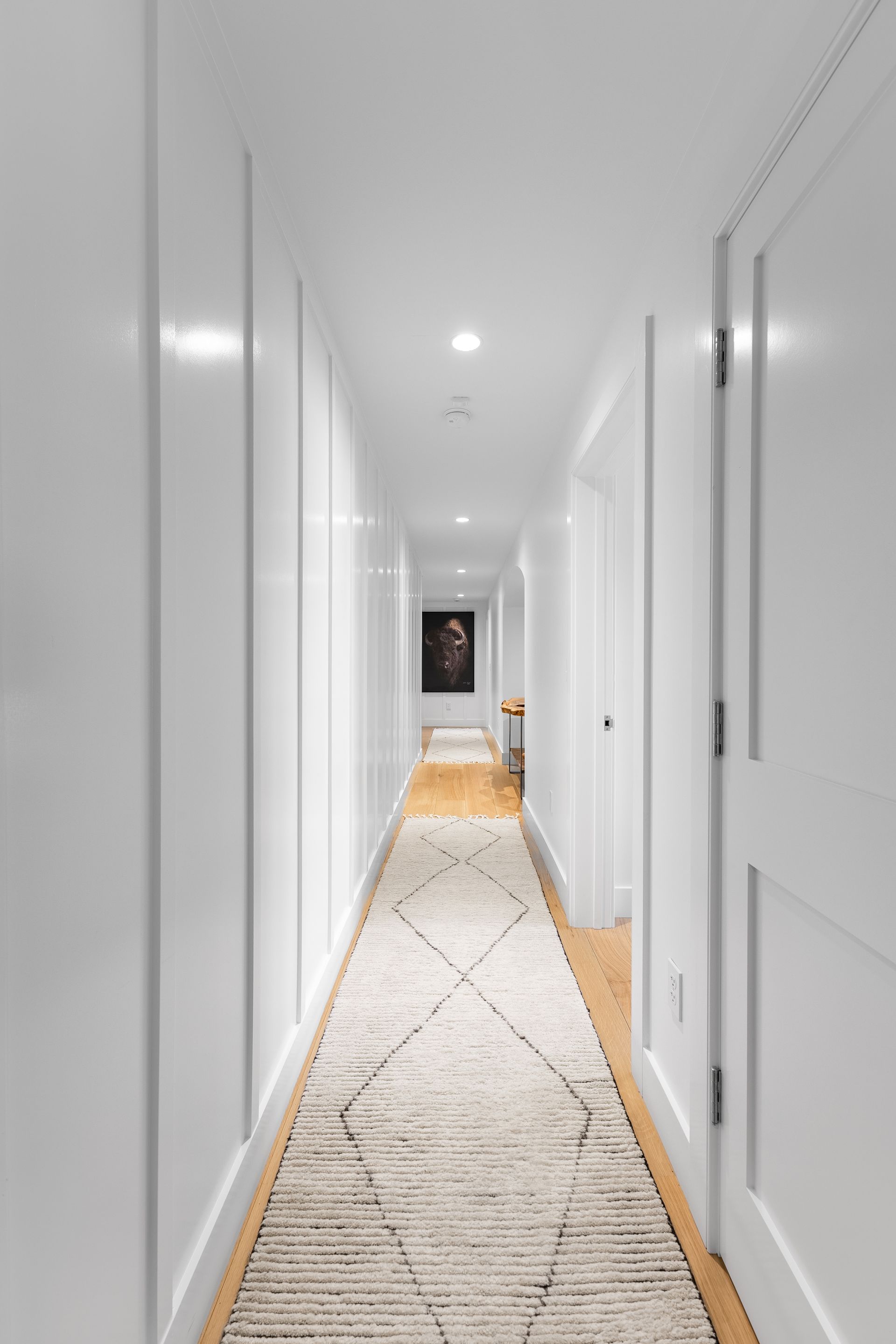 Long white hallway with doors and a patterned runner leading to a framed artwork at the far end
