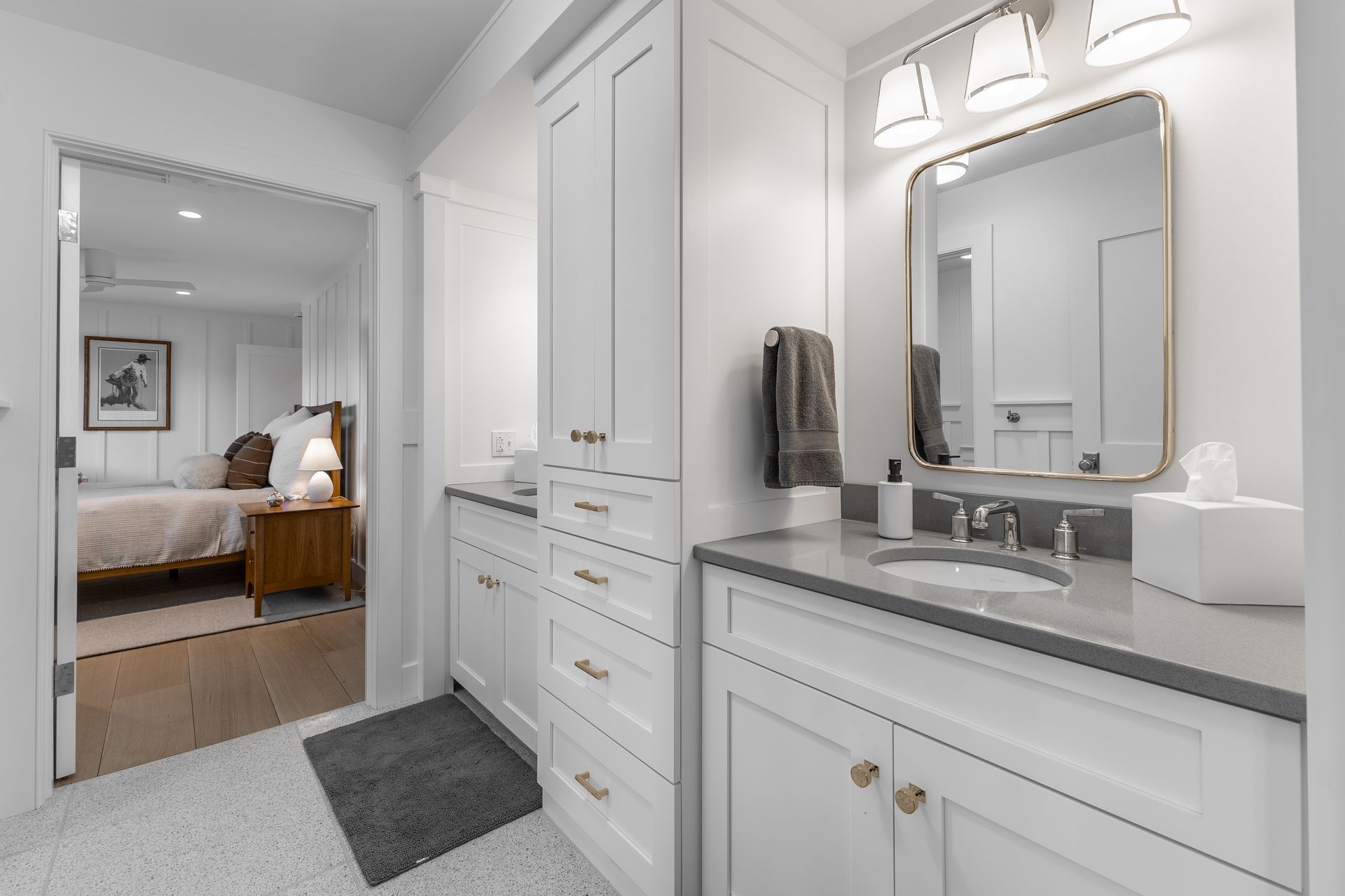 Bright white bathroom with double vanity, gold-trimmed mirror, and view into a bedroom.