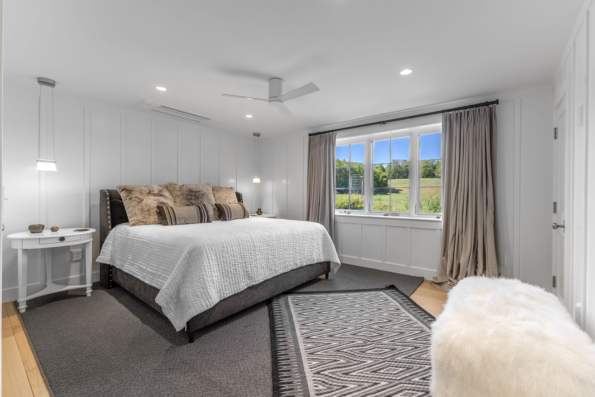 Bright bedroom with a bed, patterned rug, white walls, and a window overlooking greenery.