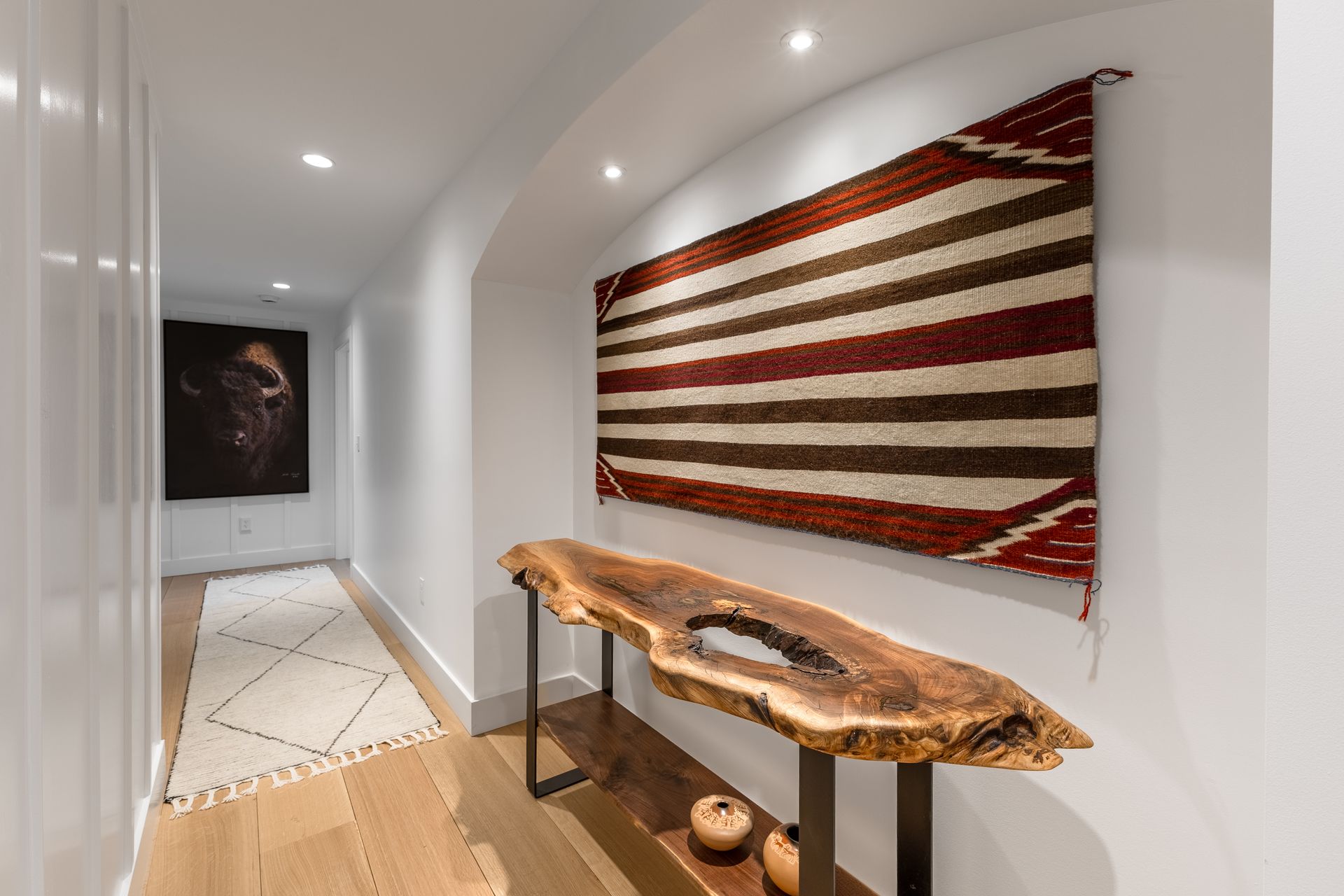 Modern hallway with white walls, wood floor, woven wall hanging, and a long rustic table with art objects
