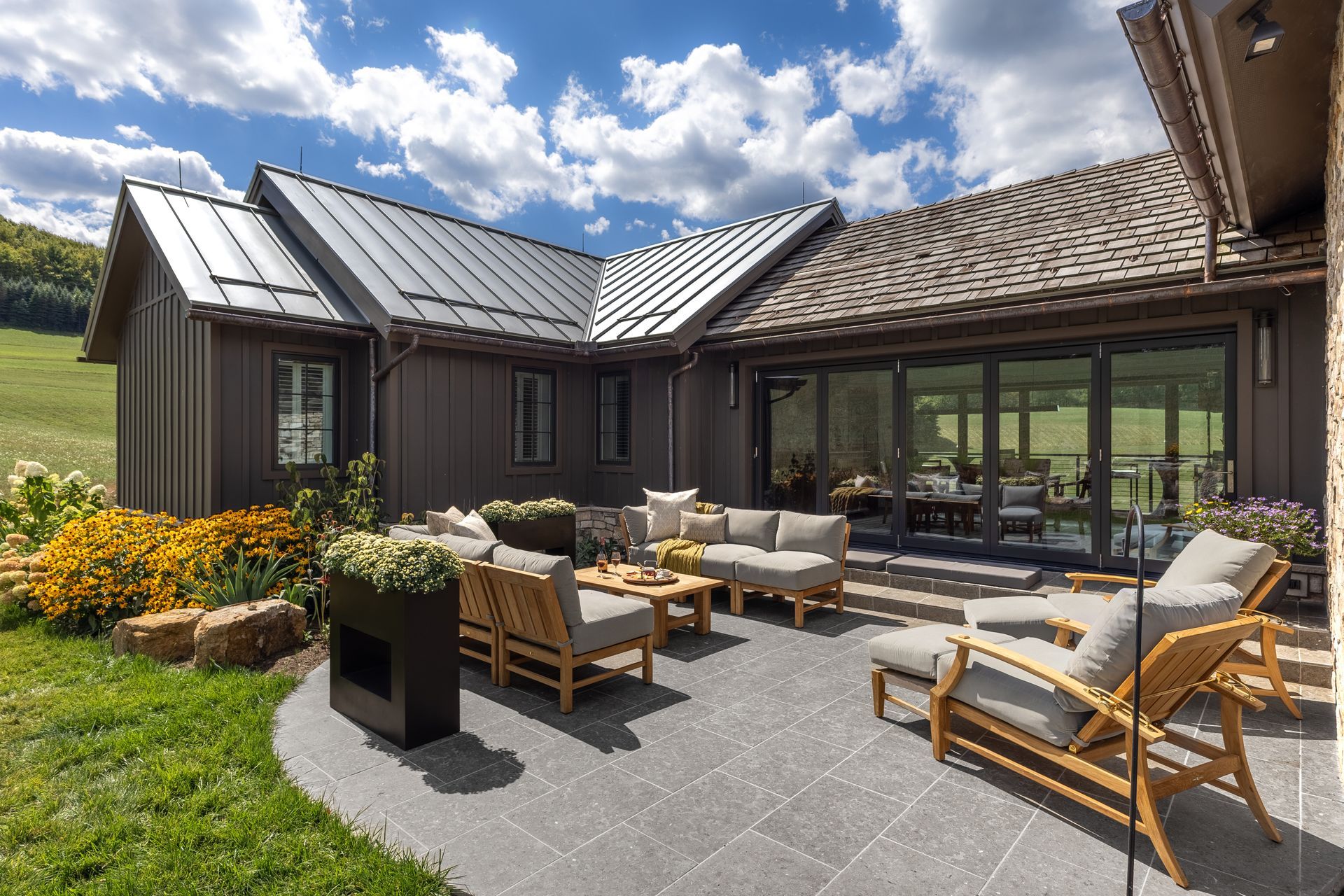 Modern patio with outdoor seating beside a dark wood house and large windows under a partly cloudy sky