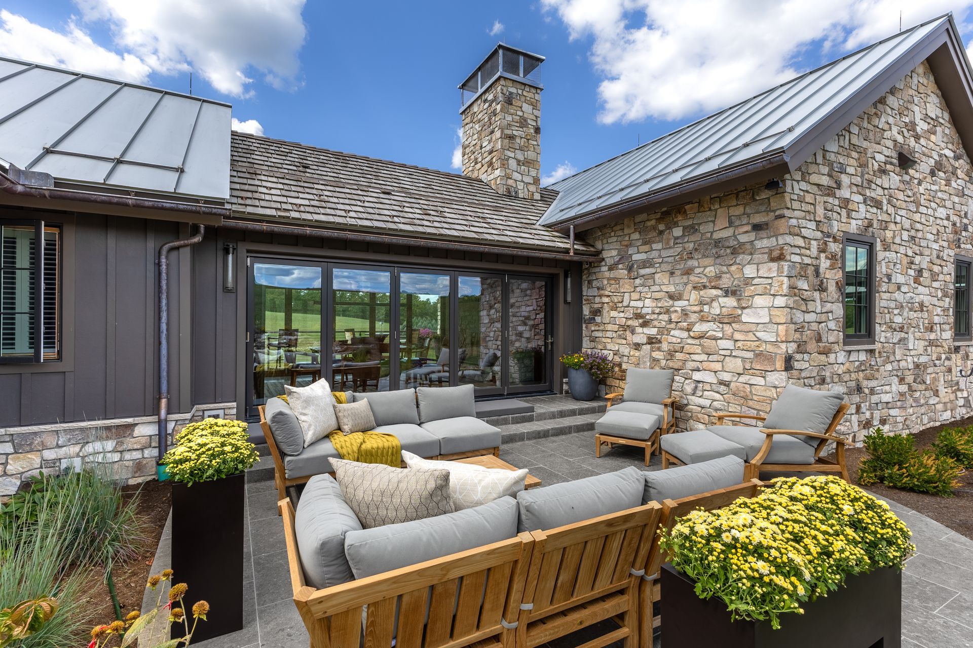 Stone patio with wicker seating, yellow flowers, and a rustic stone-and-wood house under a blue sky