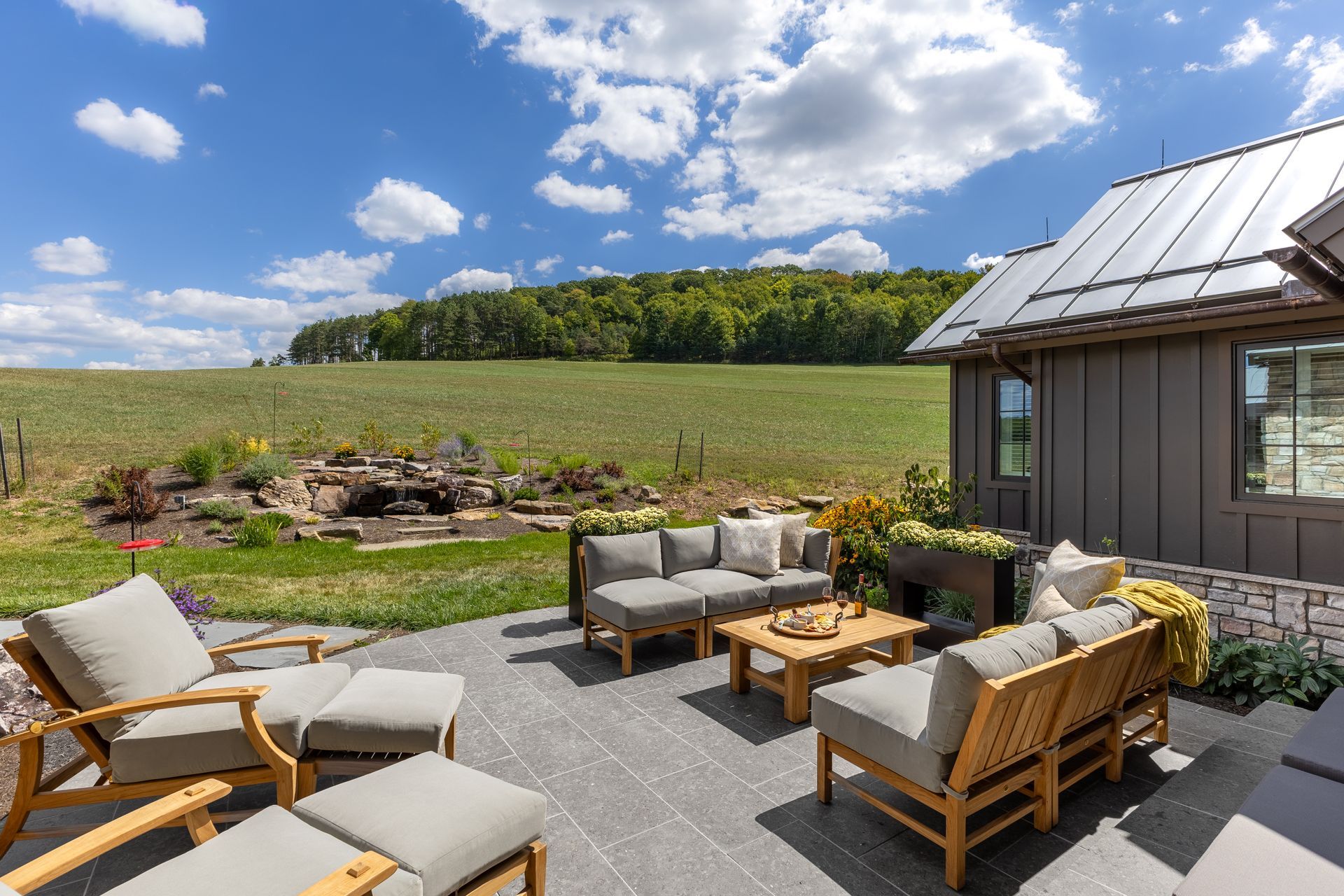 Patio with wicker chairs and table beside a rustic house, overlooking green fields under a blue sky with clouds