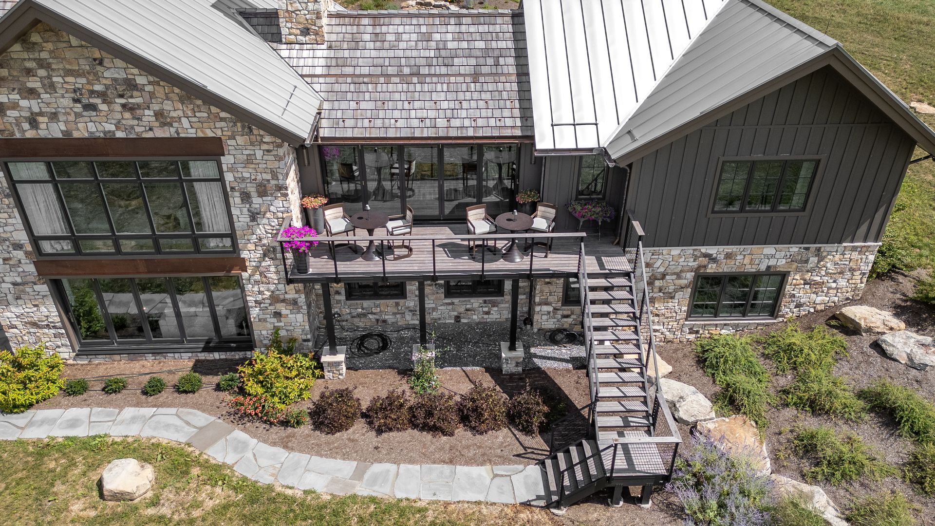 Aerial view of a stone house with a deck, outdoor seating, stairs, and landscaped garden