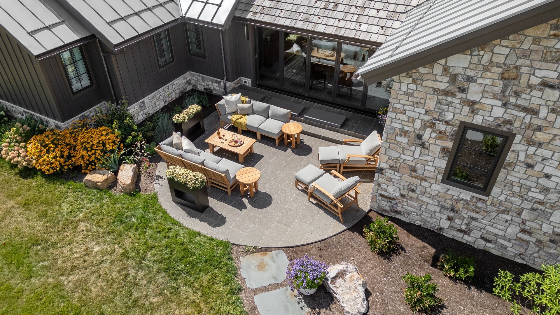 Modern patio with wicker seating, fire pit, stone house exterior, and flower beds in a landscaped backyard