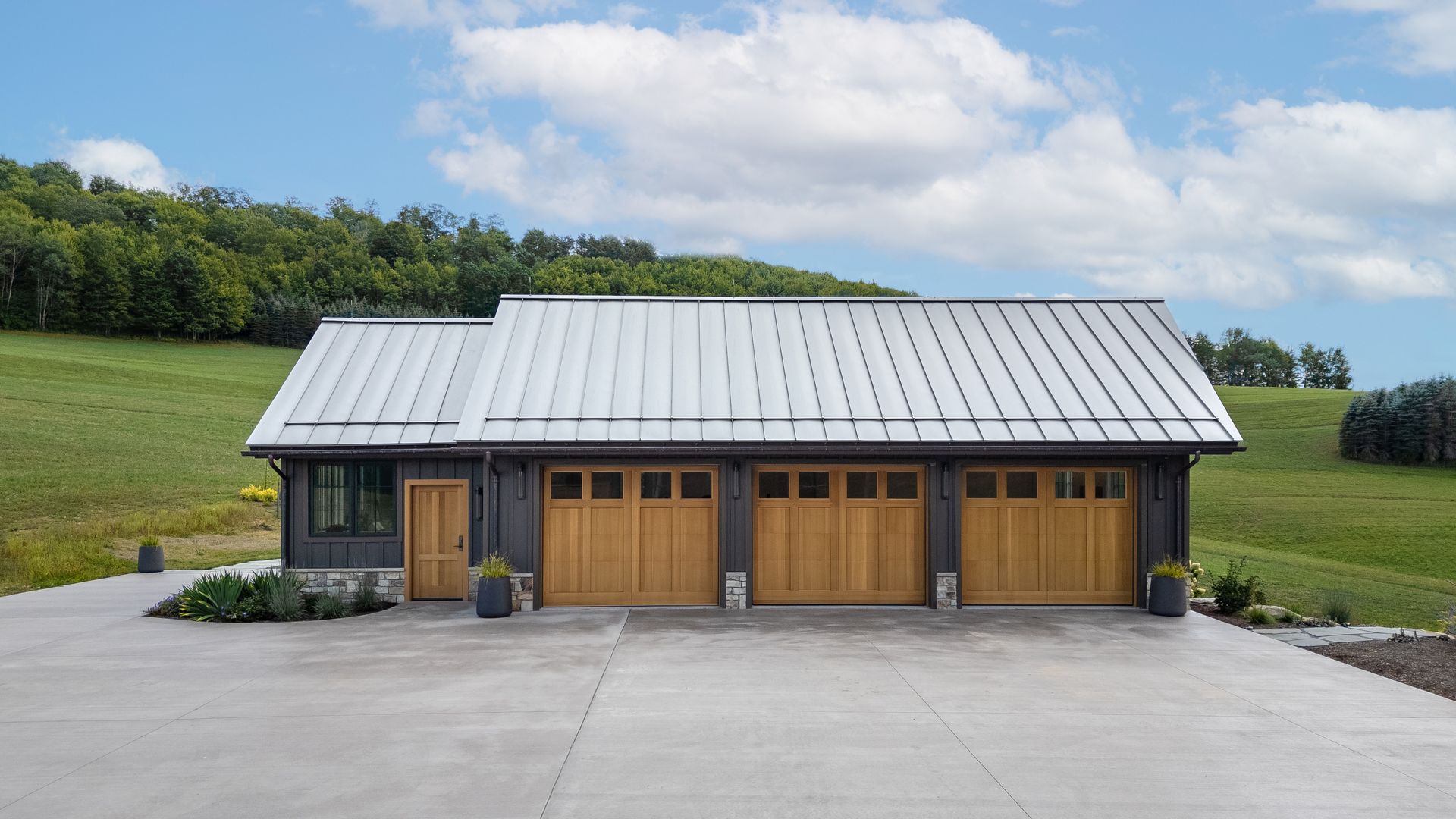 Modern gray-roofed farmhouse with three wooden garage doors on a concrete driveway, surrounded by green fields