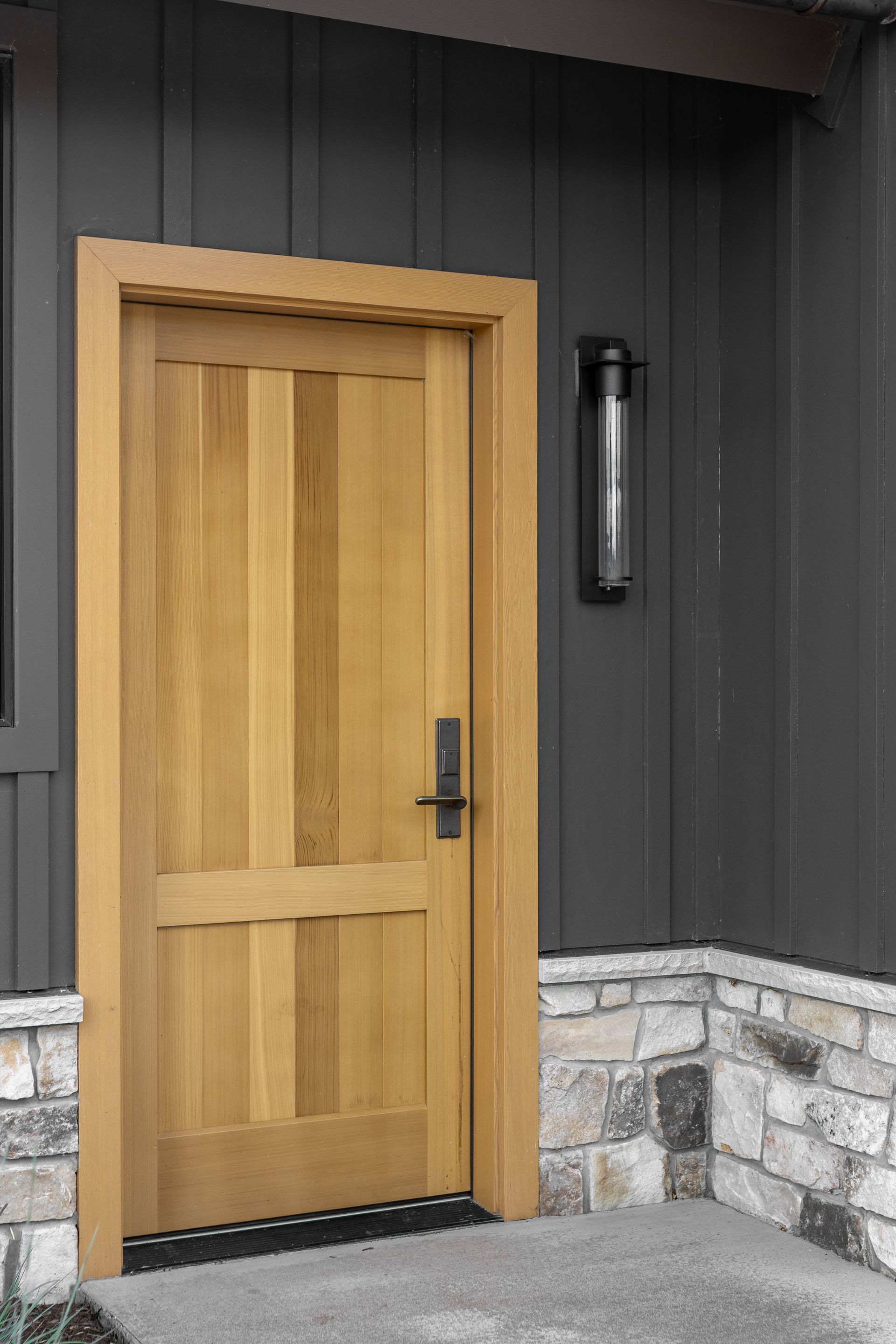 Wooden front door with black handle, set in a dark exterior wall with stone base trim