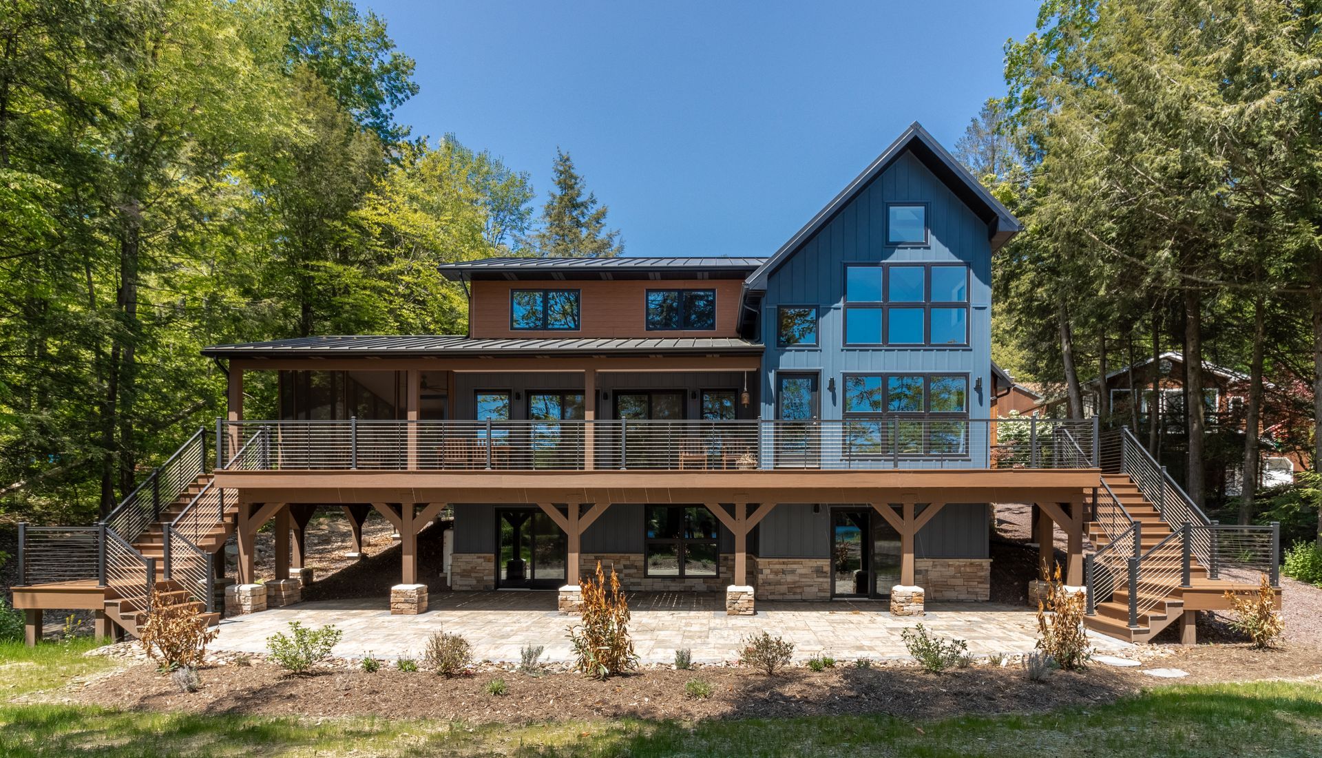 Modern house with a large deck, surrounded by trees. Blue and brown exterior, large windows.