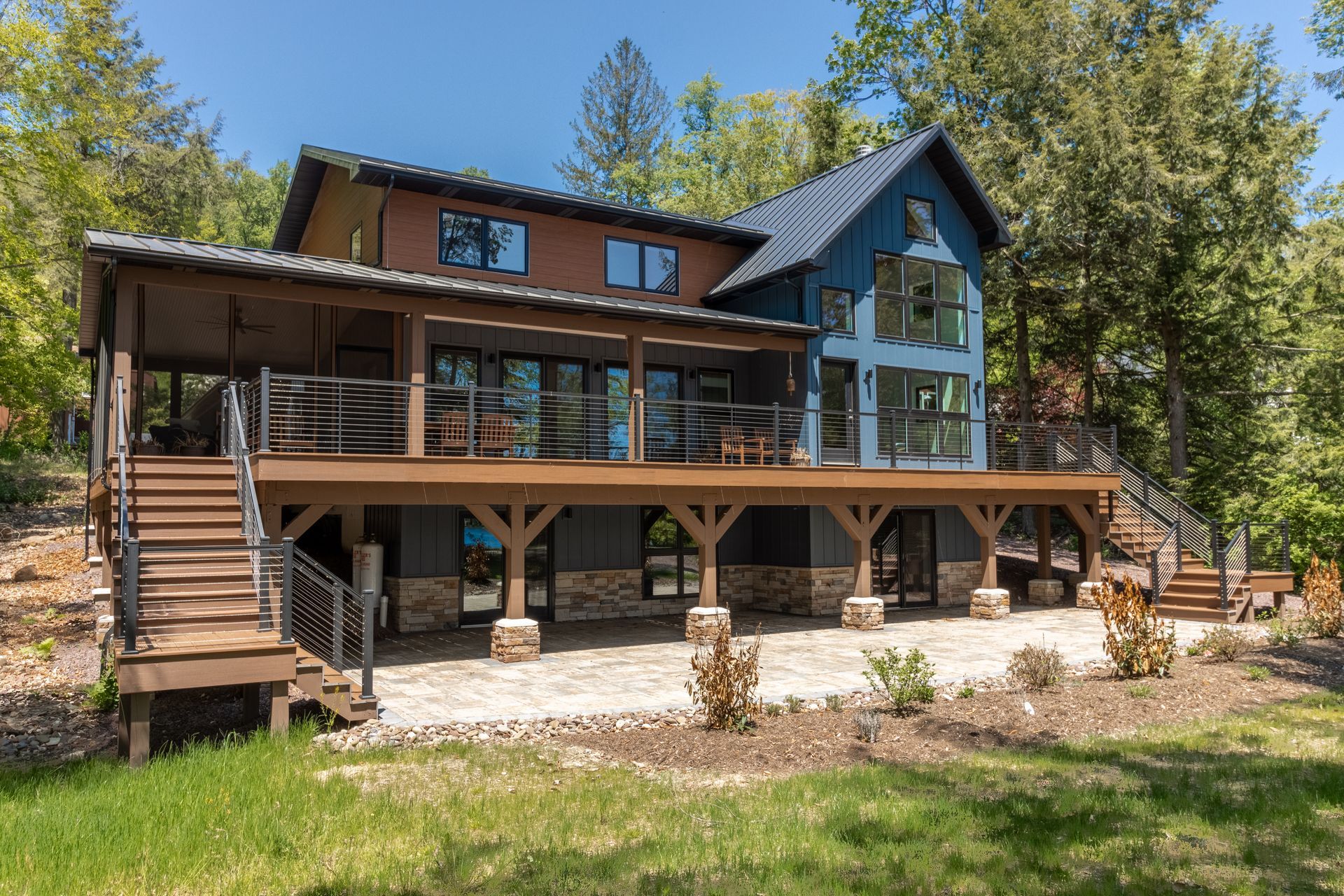 A two-story home with blue and brick siding, a large wooden deck, and a stone patio surrounded by trees.