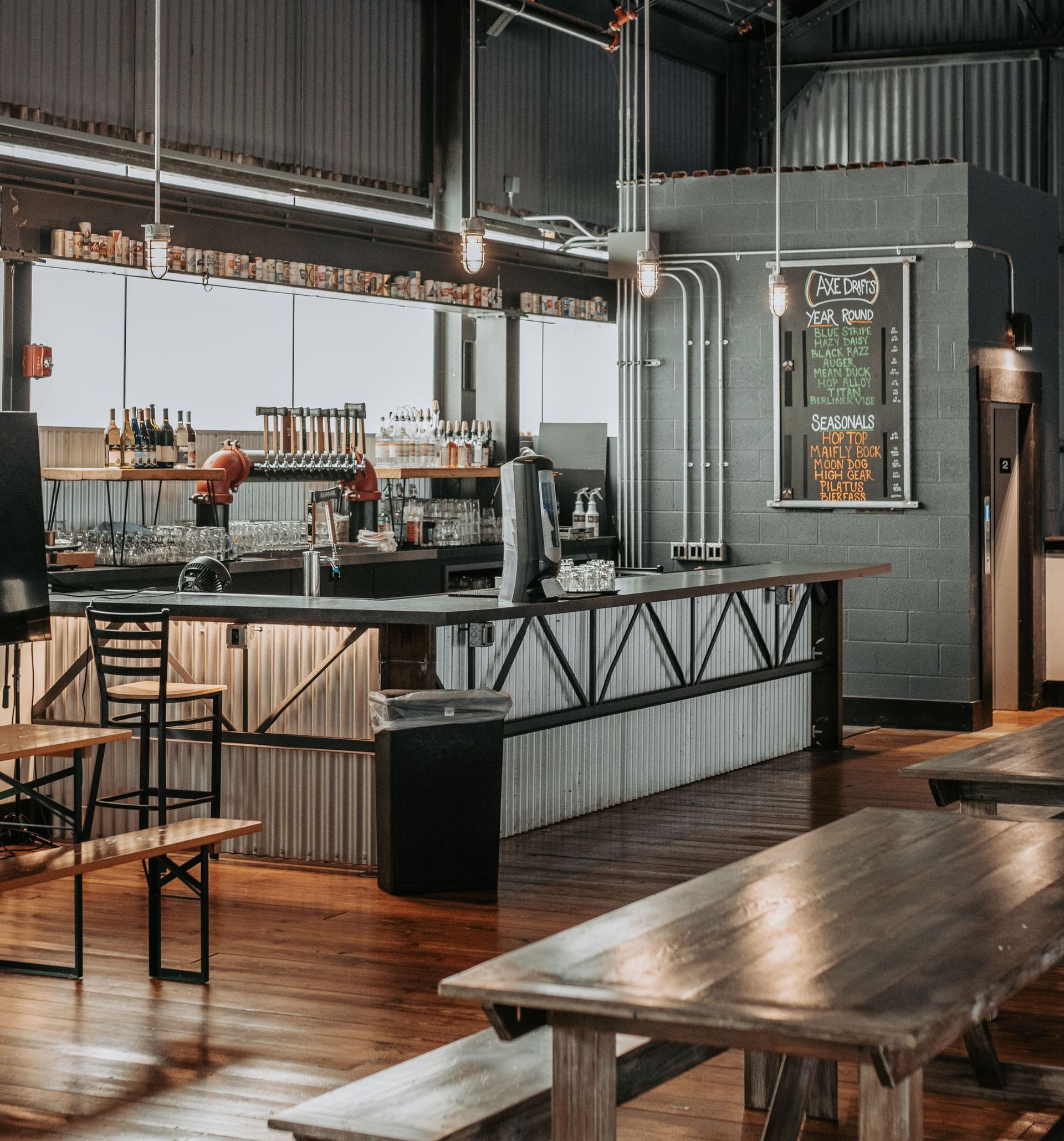 An industrial-style brewery interior with a corrugated metal bar, wooden tables, and a chalkboard menu on a gray wall.