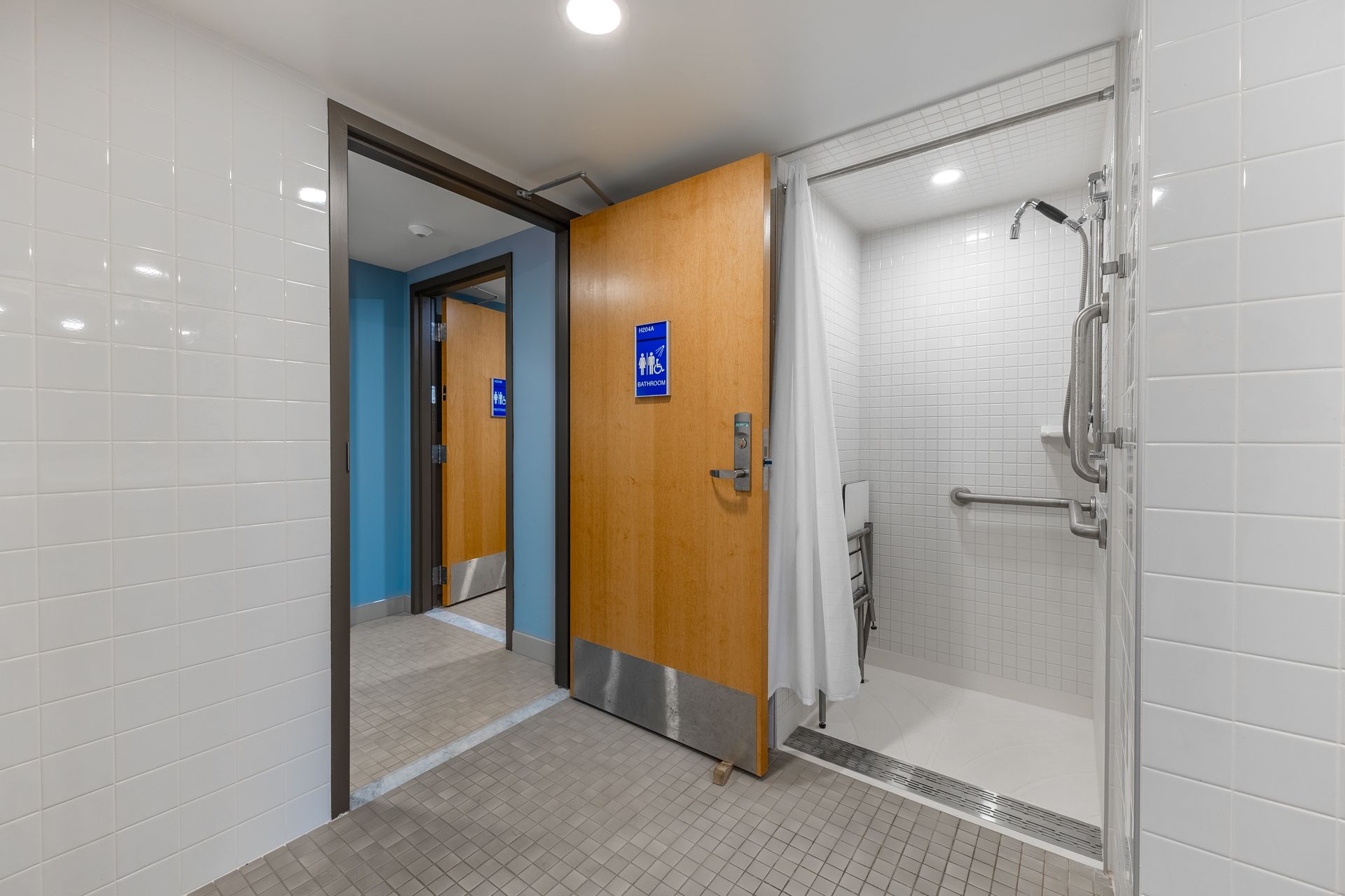 Hamilton Hall accessible bathroom entrance with wooden door, blue signs, and a tiled shower stall visible inside