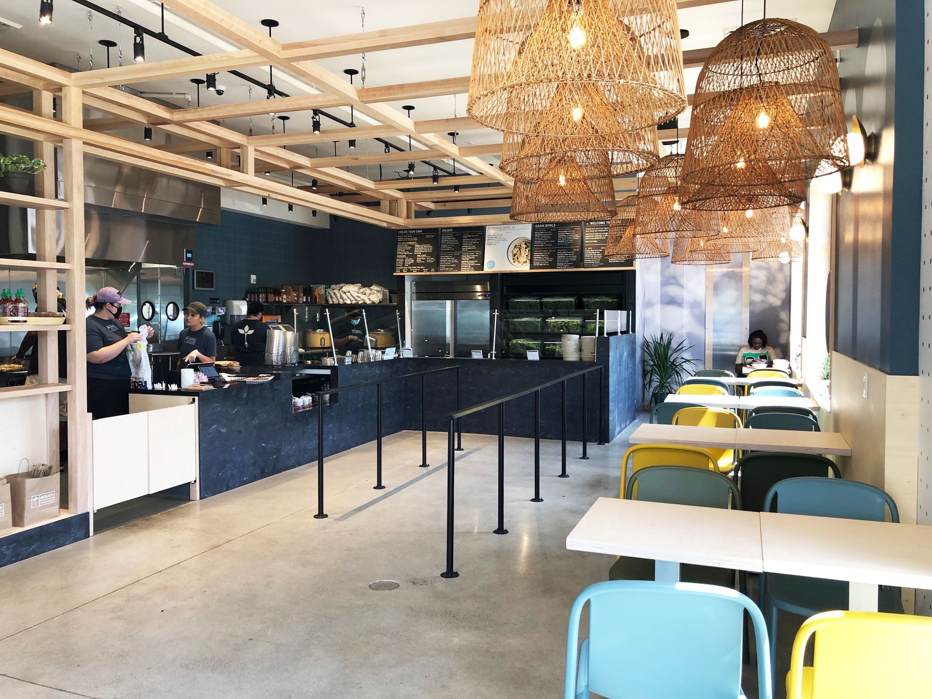 Roots Natural Kitchen interior with light wood rafters, woven pendant lights, a dark service counter, and colorful metal chairs.