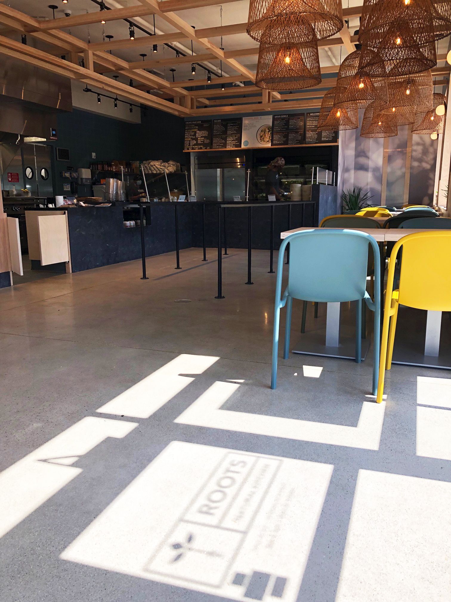 Roots Natural Kitchen interior with blue and yellow chairs, dark bar, and sunlight streaming through windows.