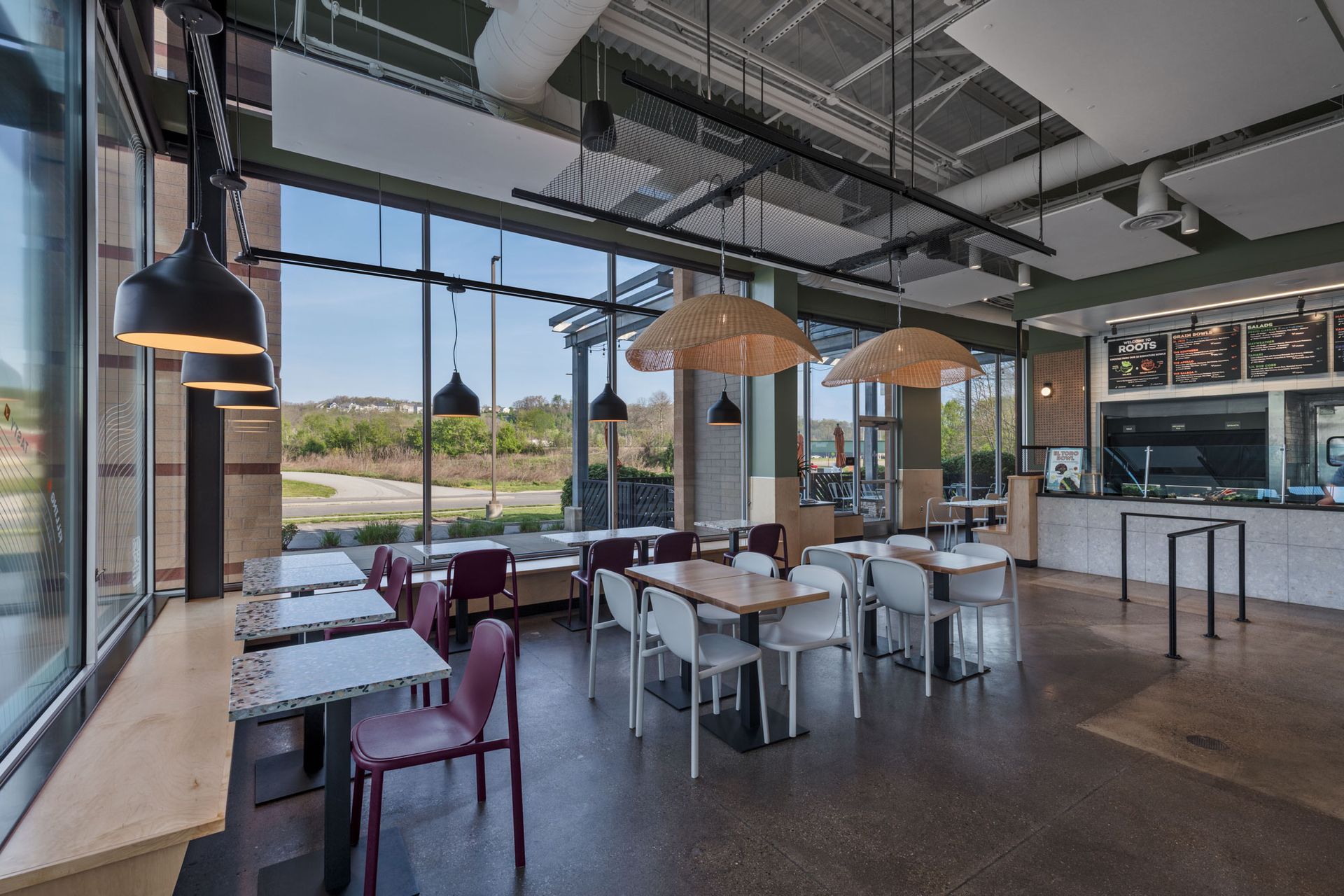 Roots Natural Kitchen's interior with white tables, wicker pendant lights, and large windows overlooking greenery