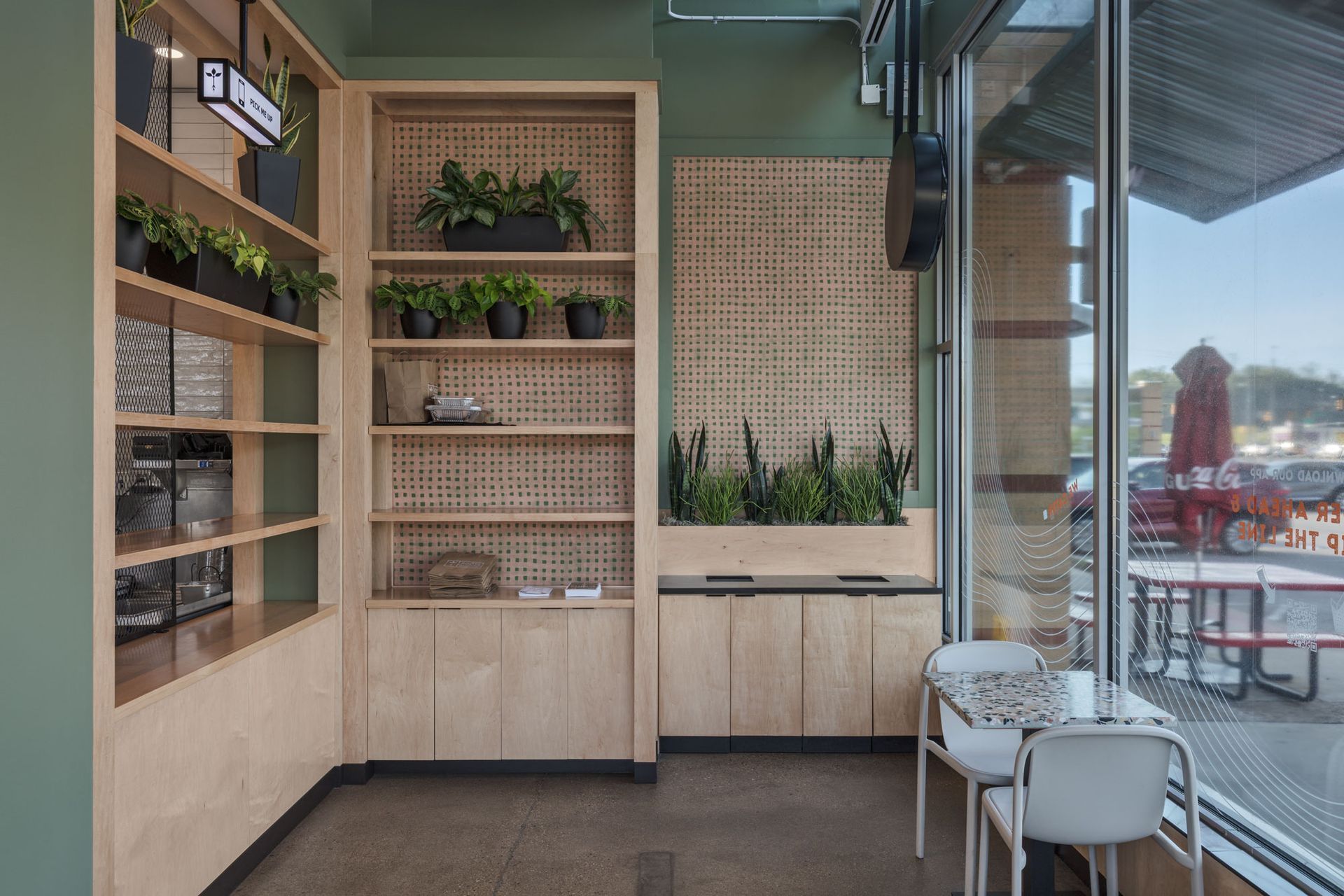 Roots Natural Kitchen's interior corner with wooden shelves, potted plants, and small tables by a glass window