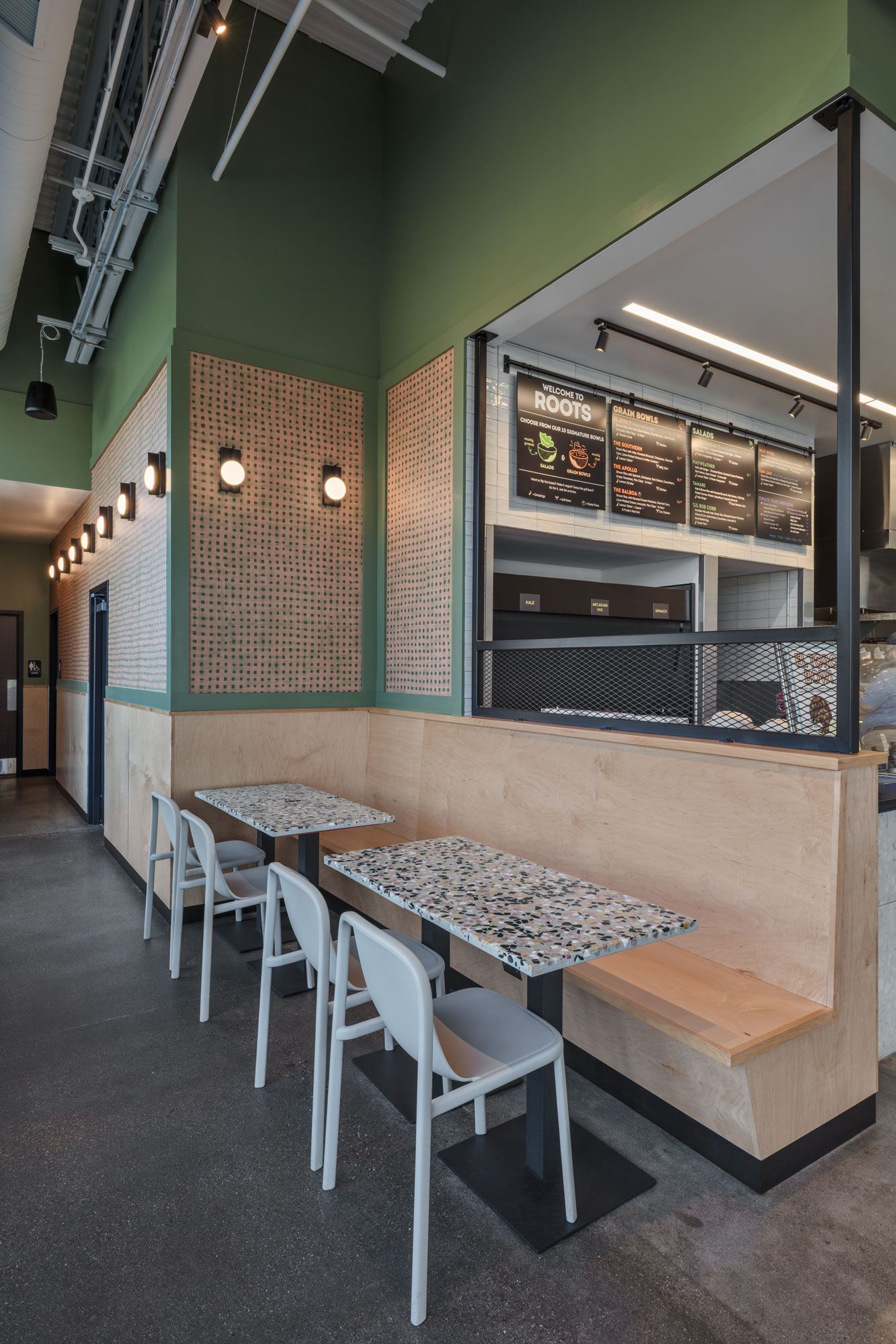 Roots Natural Kitchen's interior seating with white chairs, marble-topped tables, green walls, and a large open service window