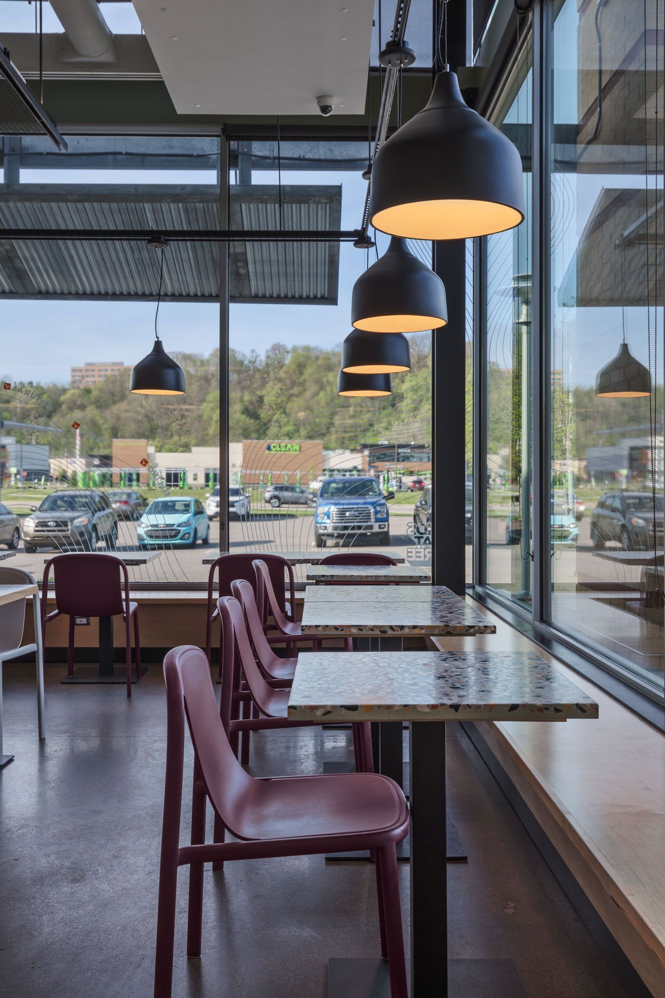 Roots Natural Kitchen's interior with pink chairs, marble tables, black pendant lights, and large windows overlooking a parking lot.