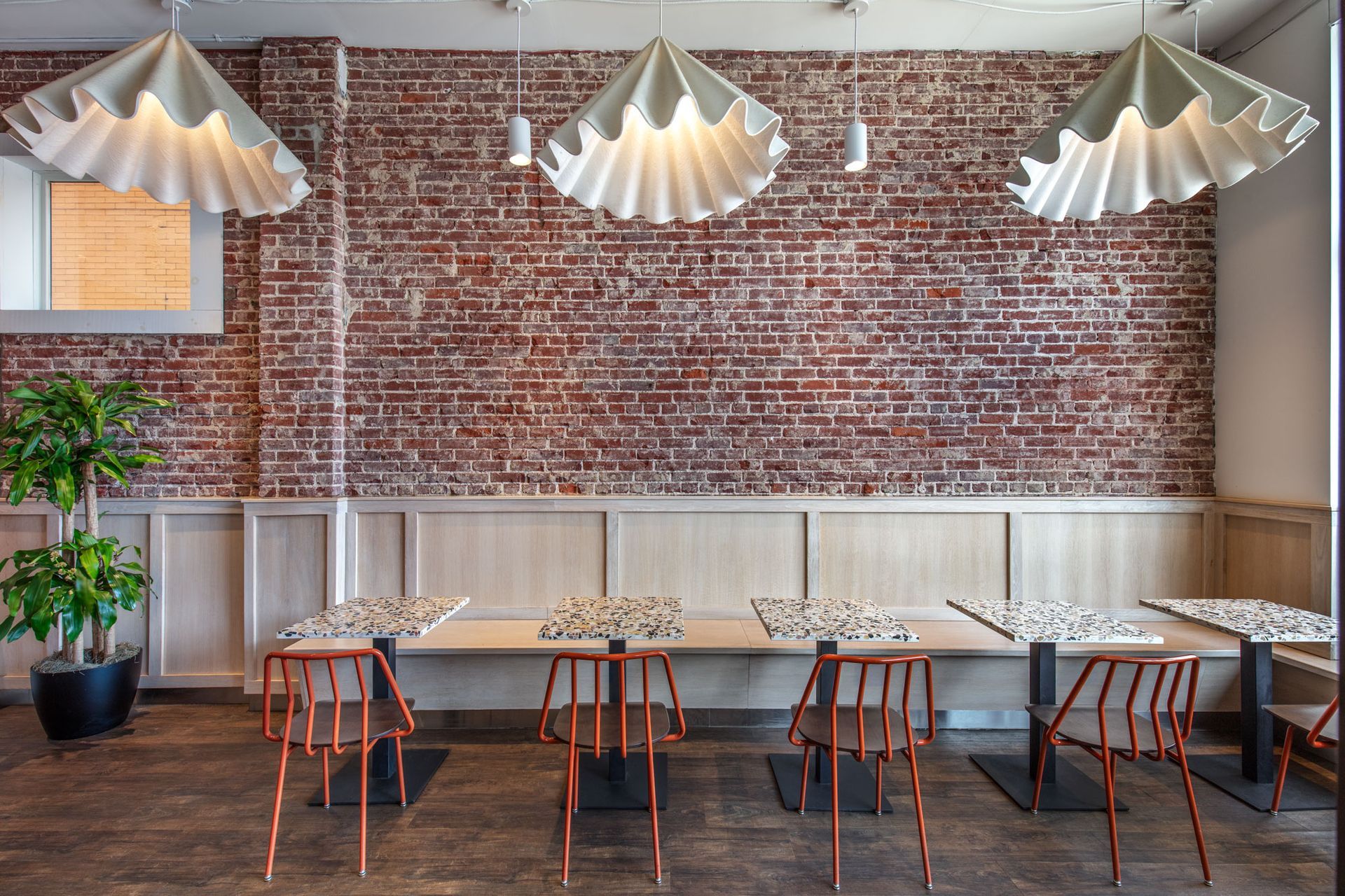Roots Natural Kitchen's interior with brick wall, hanging pendant lights, white bench, and small tables with orange chairs