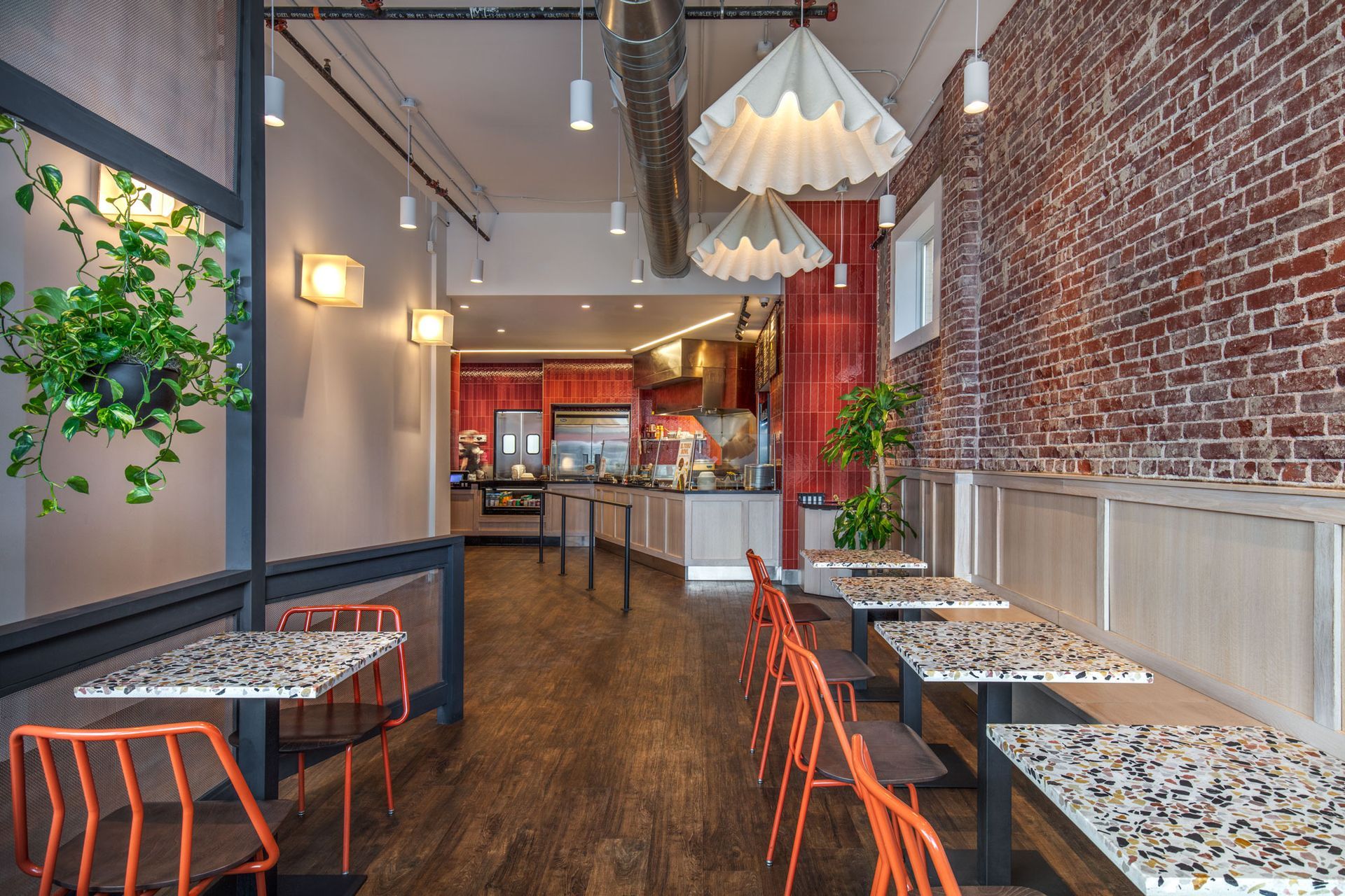 Roots Natural Kitchen's interior with brick walls, hanging chandeliers, and small tables with orange chairs on a wooden floor