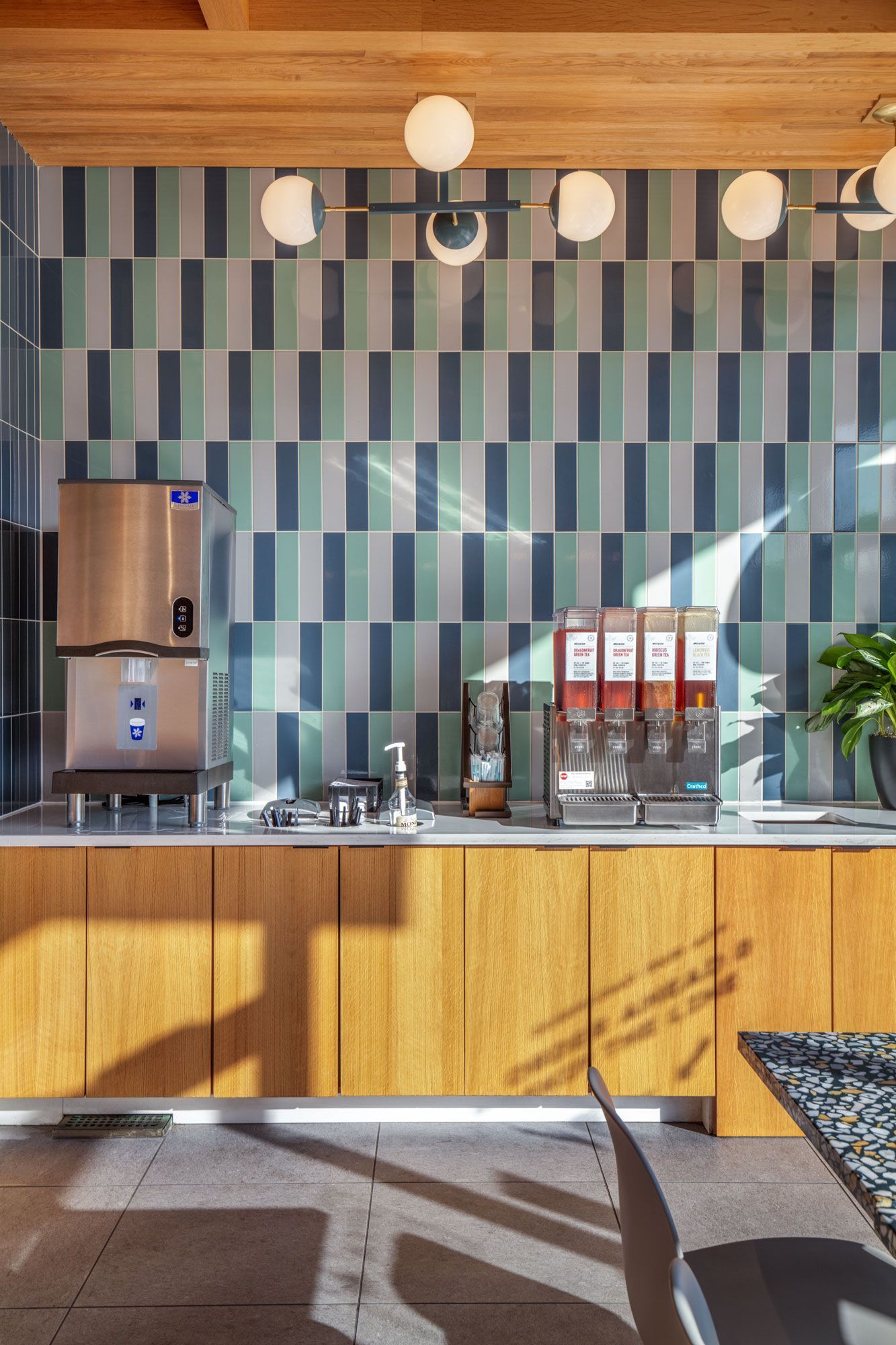Roots Natural Kitchen's interior counter with striped tile wall, wooden cabinets, pendant lights, and sunlit seating area.