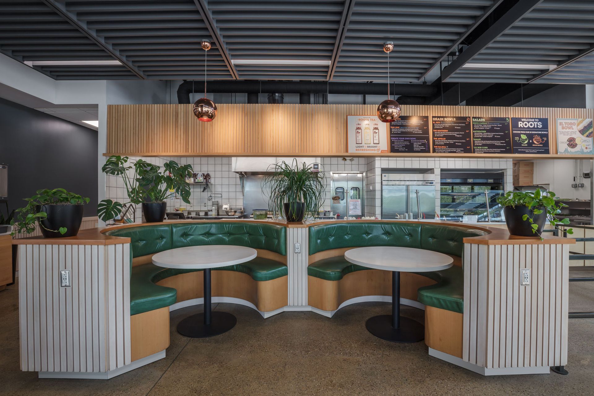 Roots Natural Kitchen's interior seating area with green booths, round tables, wood paneling, and menu boards under industrial lighting