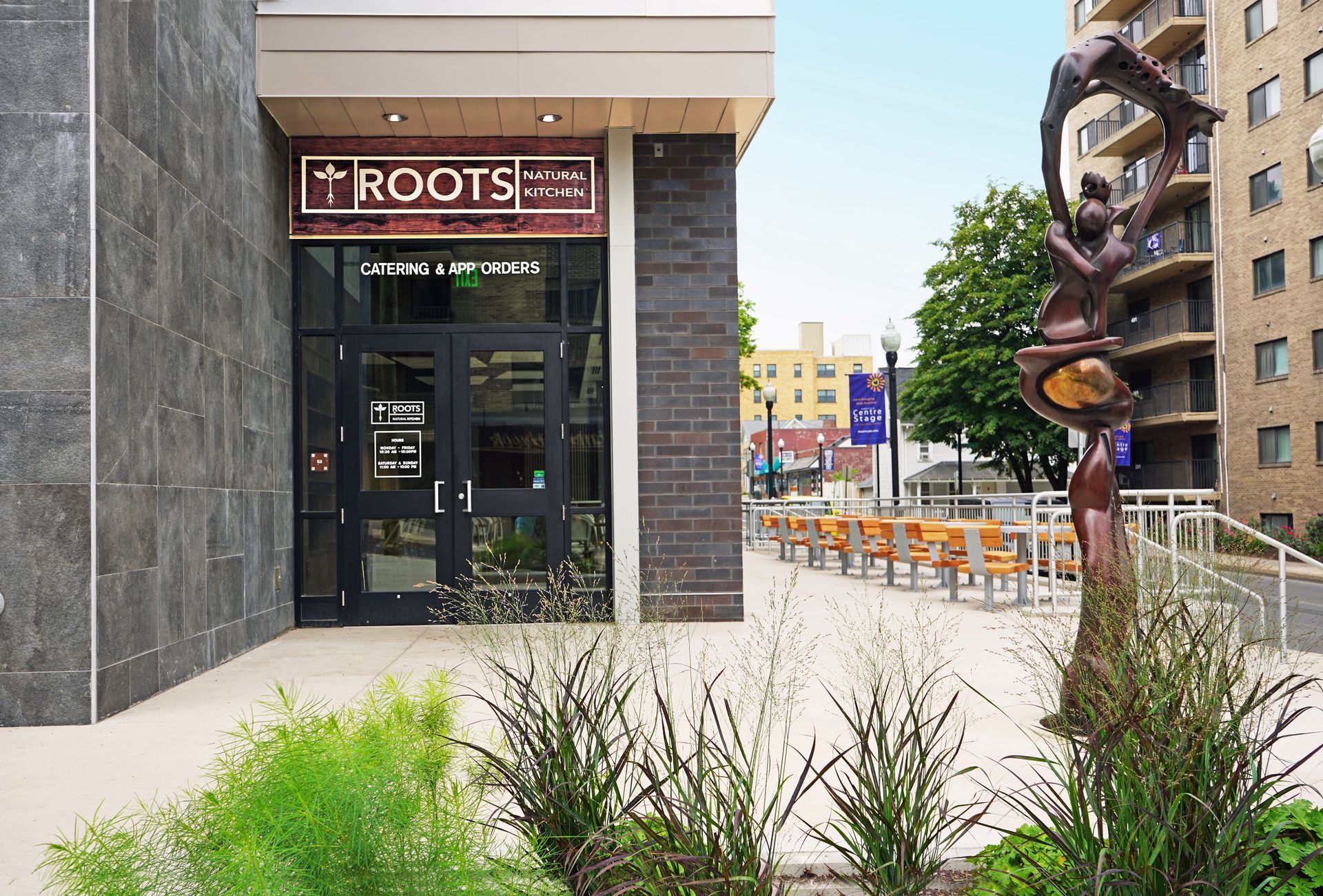 Roots Natural Kitchen's storefront entrance with “ROOTS” sign on a city sidewalk, with landscaping and a sculpture nearby