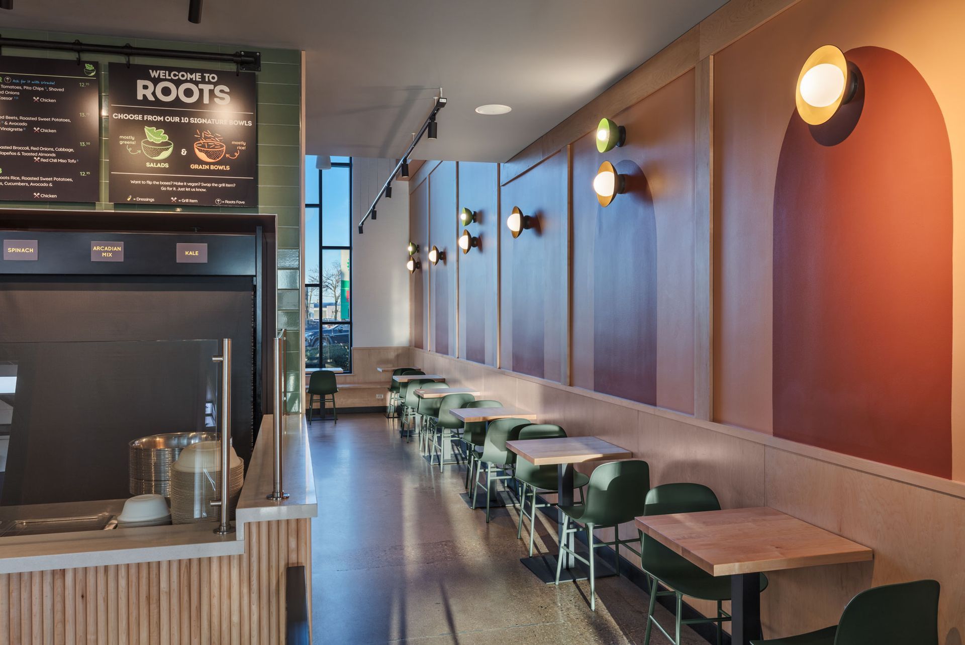 Roots Natural Kitchen's interior with long wooden tables, green chairs, and warm wall lights along a corridor-like seating area