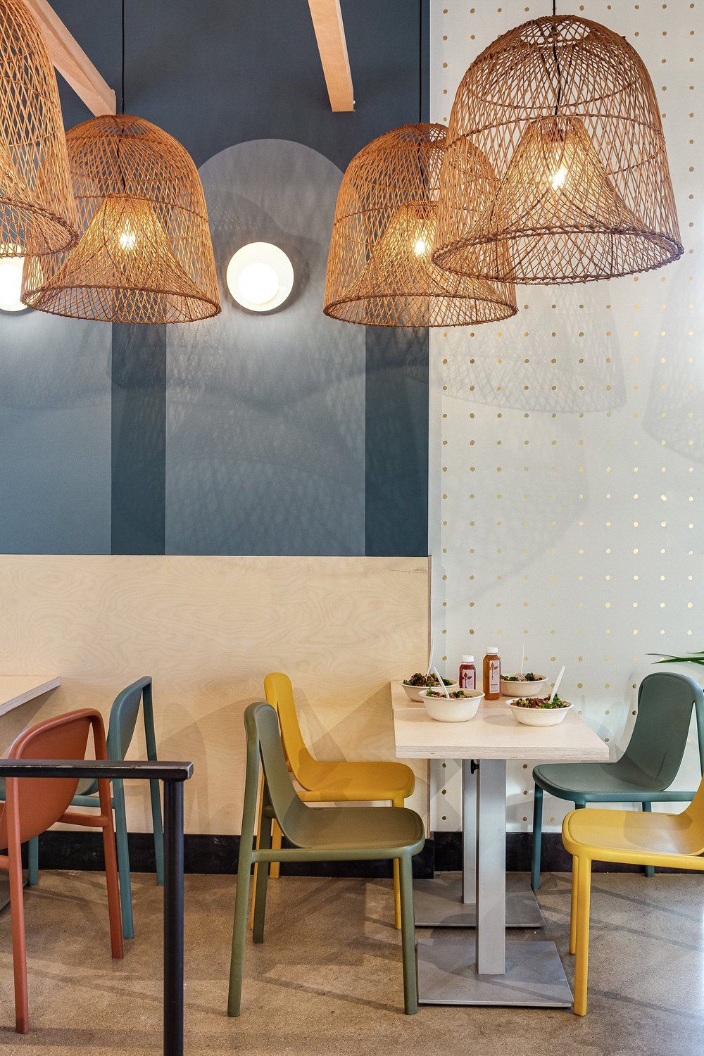 Roots Natural Kitchen interior with woven pendant lights, colorful chairs, and small tables against a blue-and-cream wall