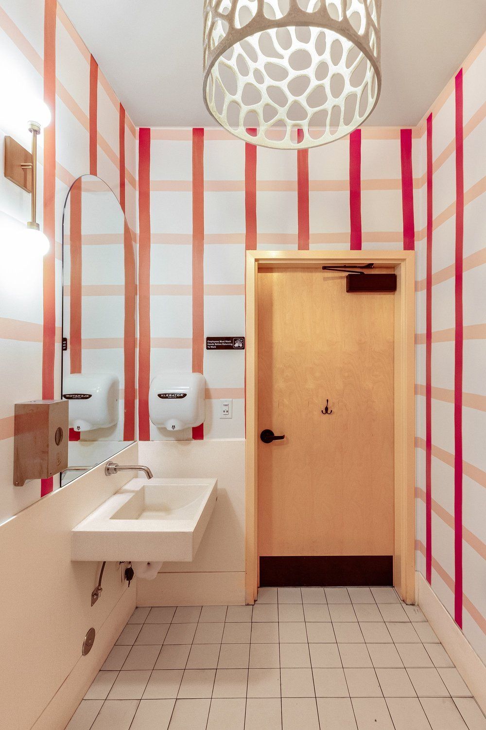 Bright narrow bathroom with white tiles, red stripes, sink, hand dryer, and wooden door.