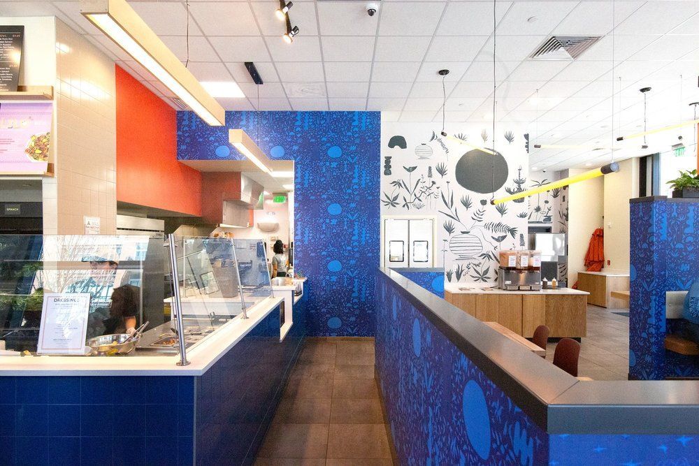 Interior of Roots Natural Kitchen with bright blue patterned walls, orange accents, a service counter, and dining area.