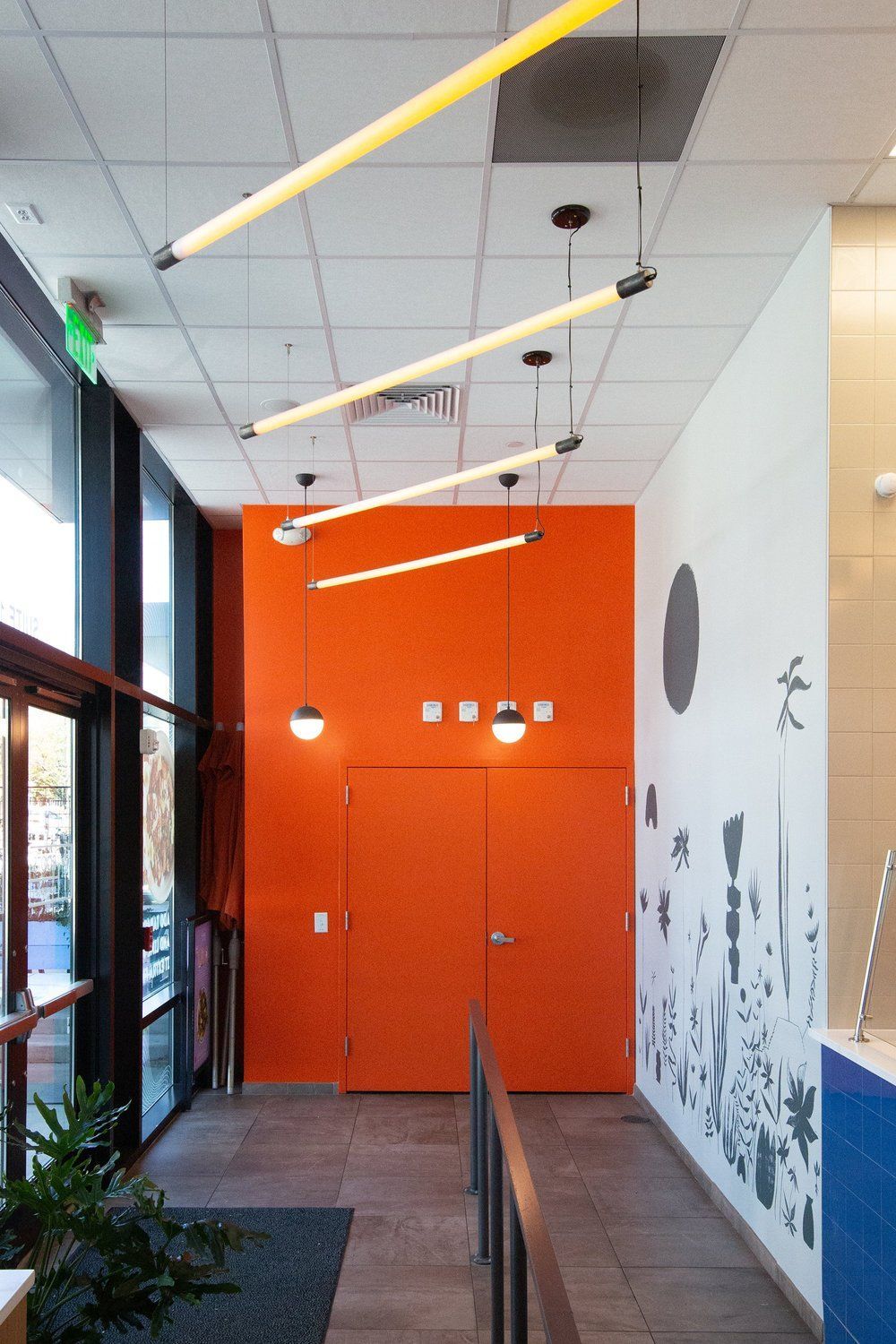 Roots Natural Kitchen's hallway with orange wall, hanging lights, glass entrance, and white wall mural