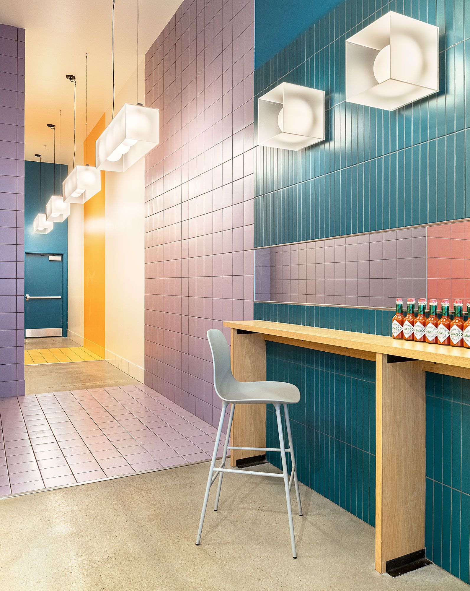 Colorful cafe interior with a bar and stools. Purple, blue, and orange walls, hexagonal lights, and a wooden counter with hot sauce.